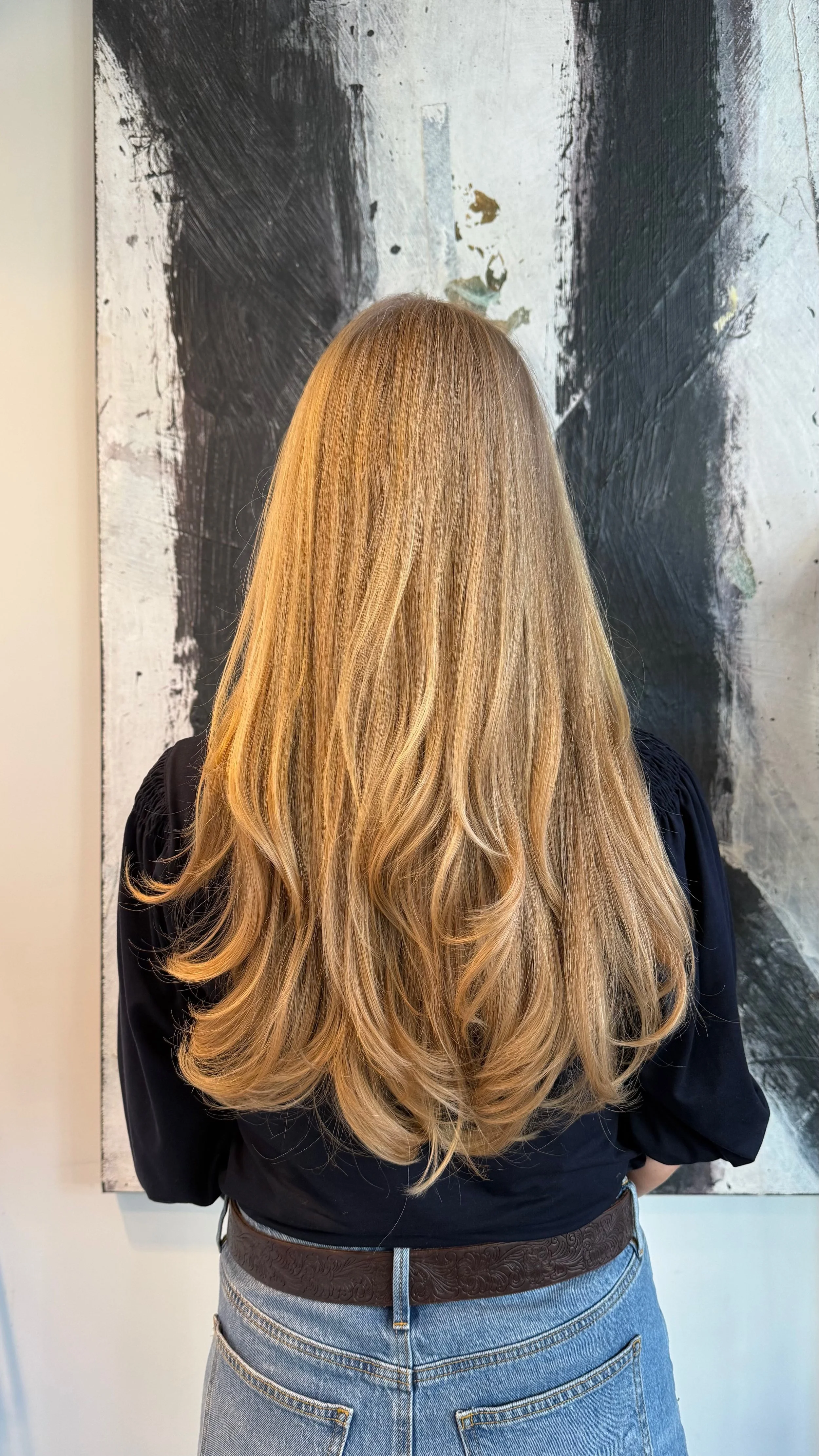 A woman with long, wavy blonde hair standing in front of an abstract black and white painting, wearing a black top and blue jeans.