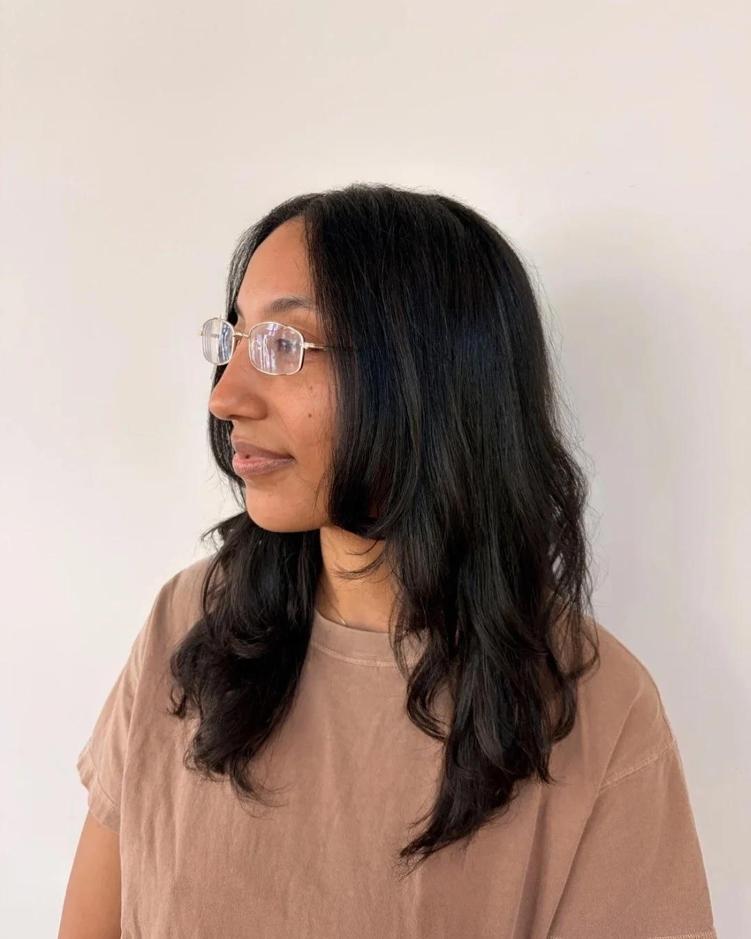 Side profile of a woman with long black wavy hair, wearing clear glasses and a beige t-shirt, standing against a plain white wall.