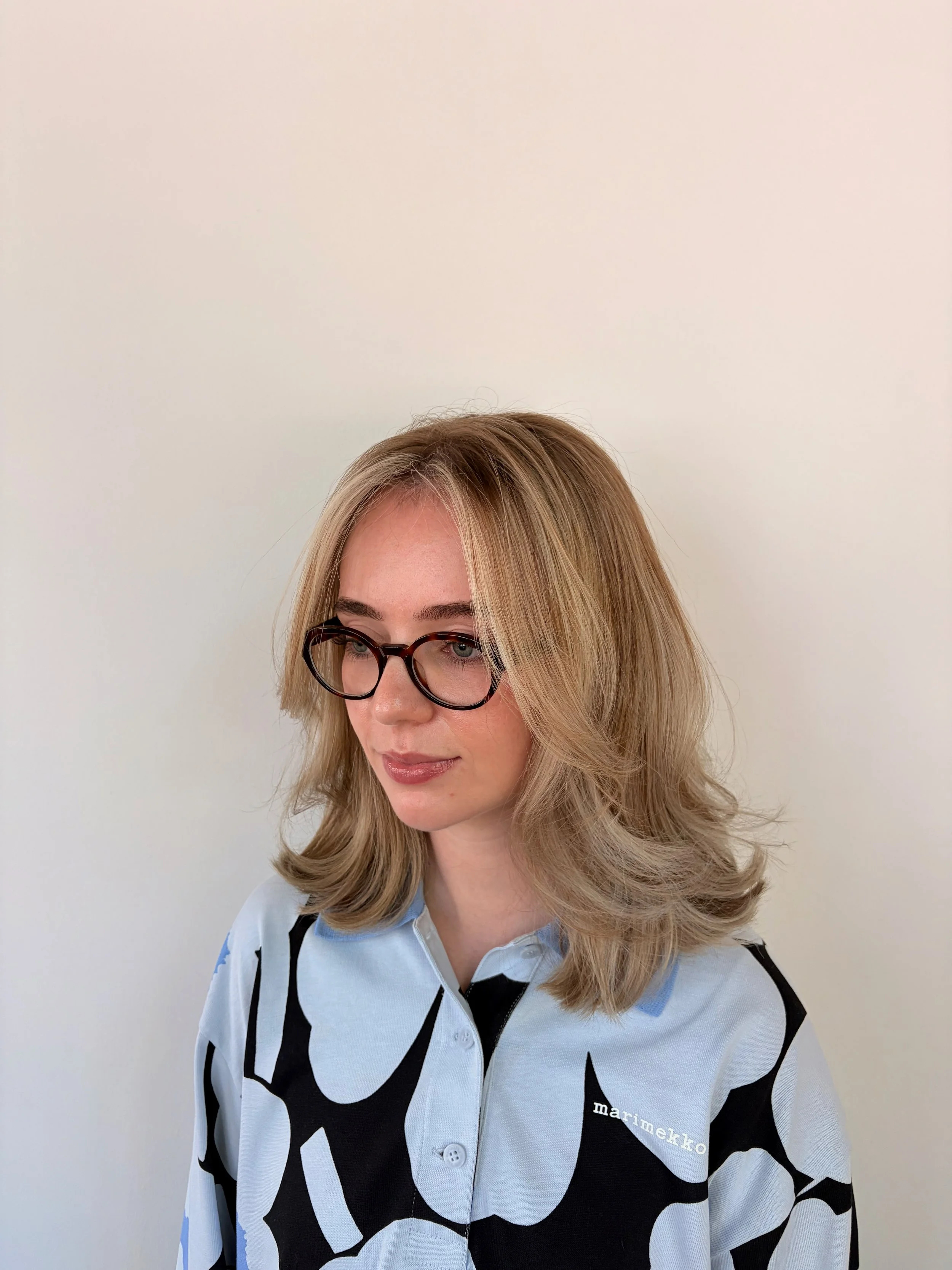 A woman with blonde hair and glasses wearing a light blue Marimekko patterned shirt with black and white bold design, standing against a plain light-colored background.