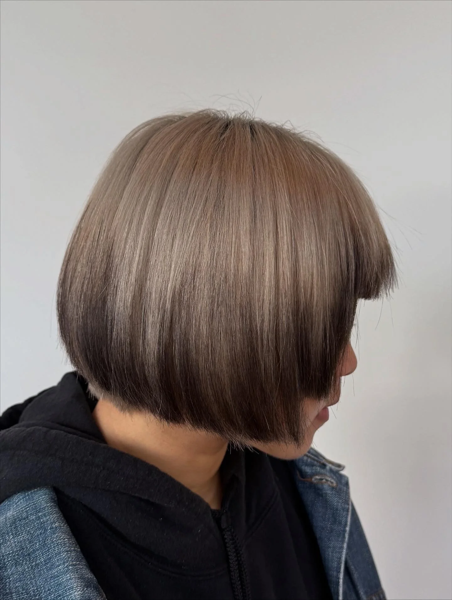 Side view of a person with a short, straight, layered haircut with shades of light brown and ash blonde, wearing a black jacket and a plaid shirt.