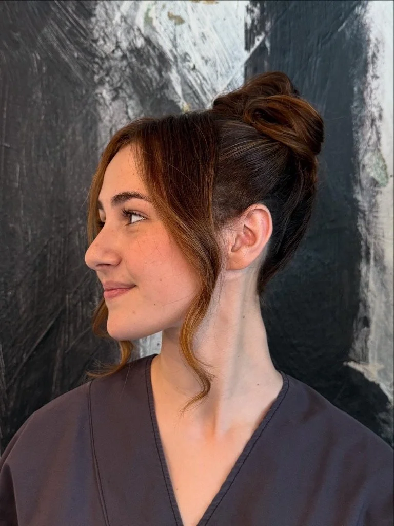 Side profile of a woman with auburn hair styled in an elevated updo, wearing a dark gray top, with a textured dark background.