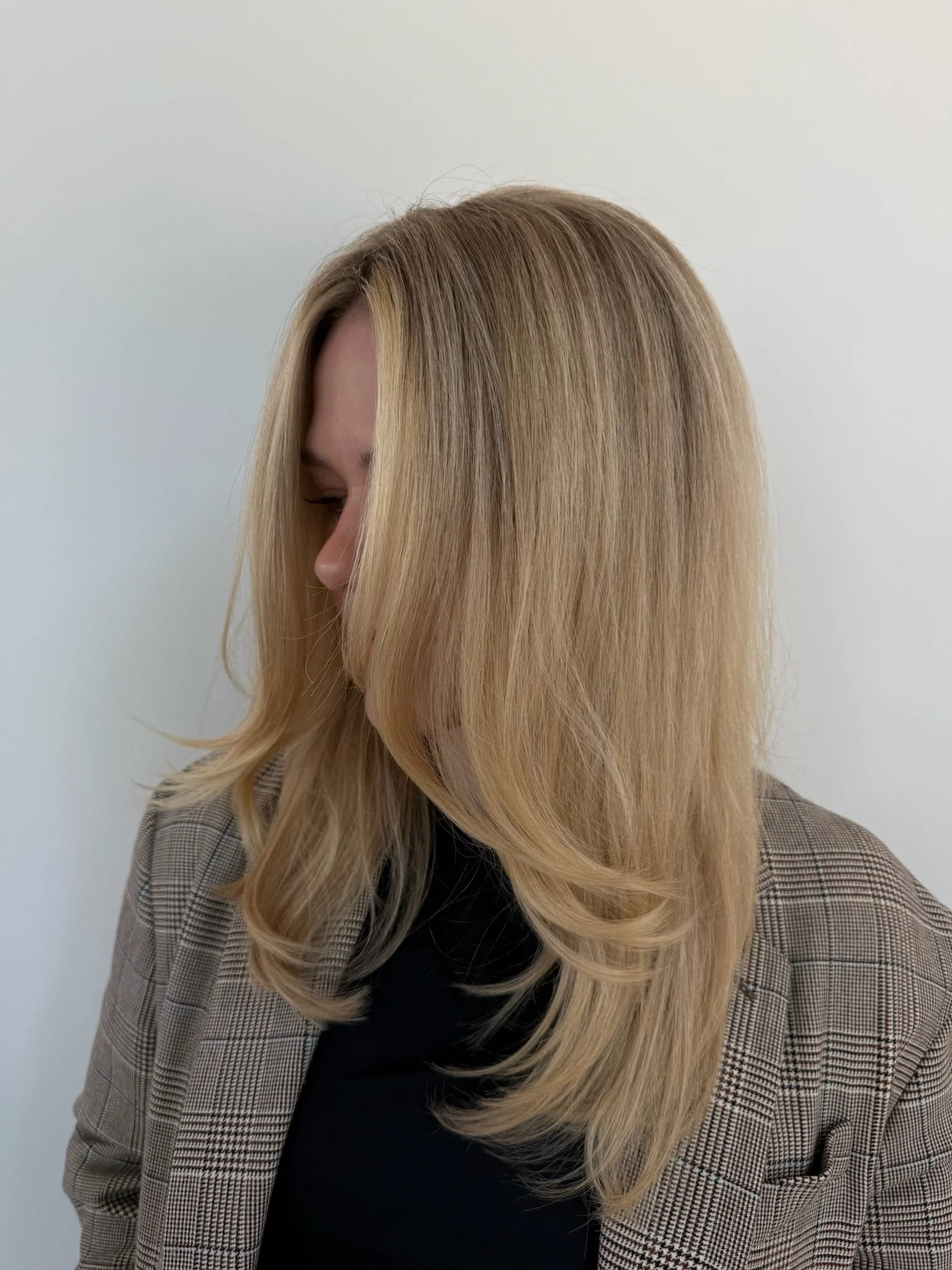A woman with long blonde hair freshly blown out, wearing a brown plaid blazer and a black top, facing slightly downward against a plain white background.