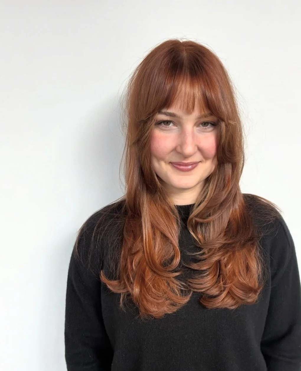 A woman with long red hair, wearing a black top, smiling and standing against a white background.