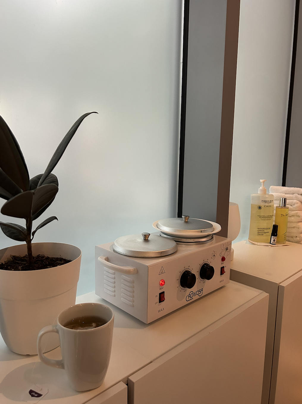 A coffee cup filled with tea, a potted plant, a wax heater with two metal lids, and bottles of hand sanitizer and lotion on a white cabinet in front of a frosted glass window.