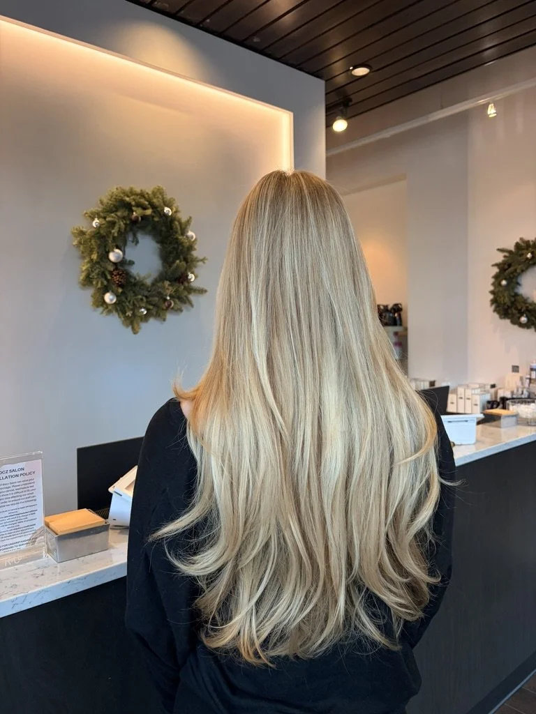 Back of a woman with long, blonde, wavy hair standing at a reception desk with holiday wreaths on the wall in a modern, well-lit interior.