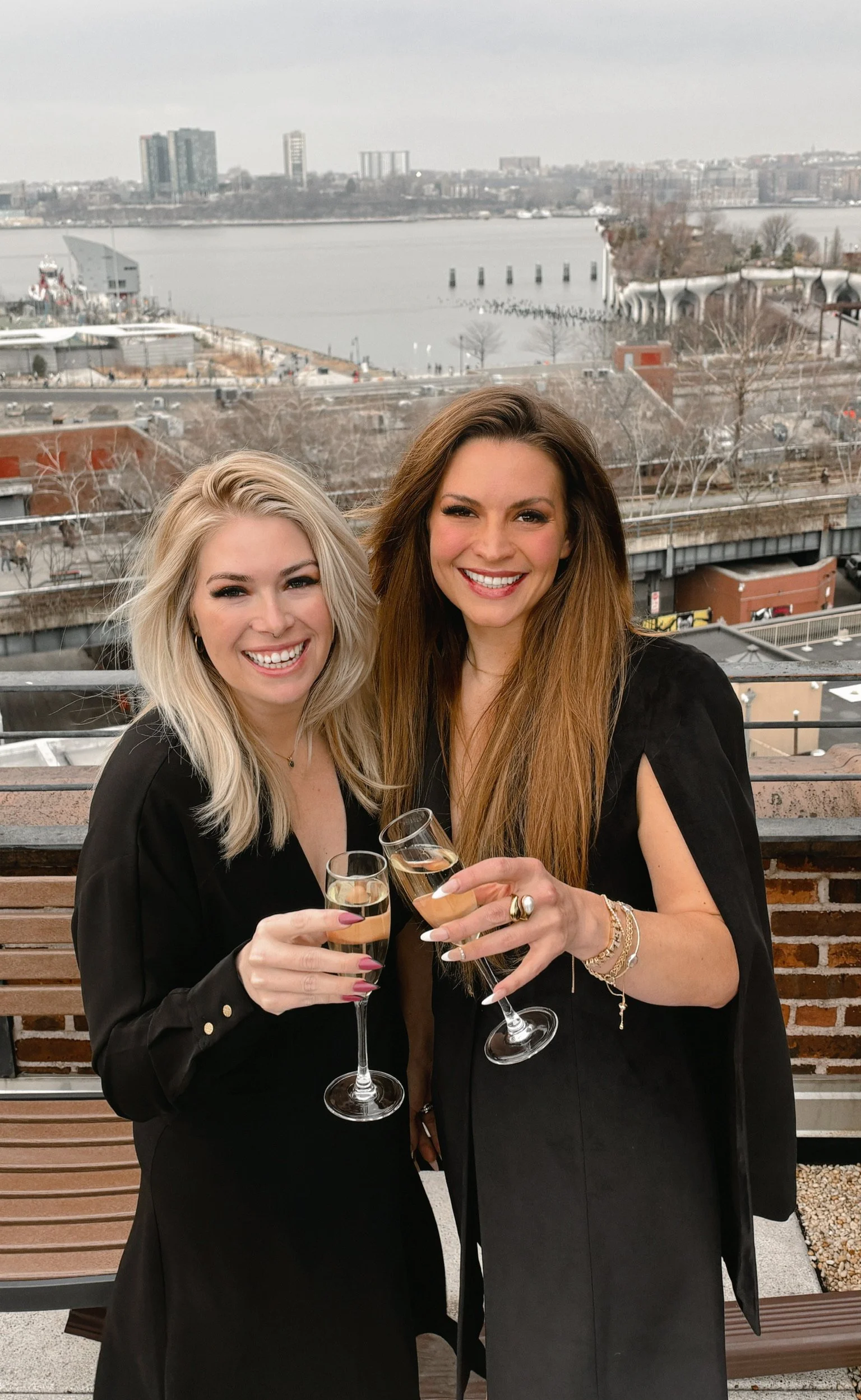 Two women smiling and clinking champagne glasses on a balcony with a city and river view in the background.