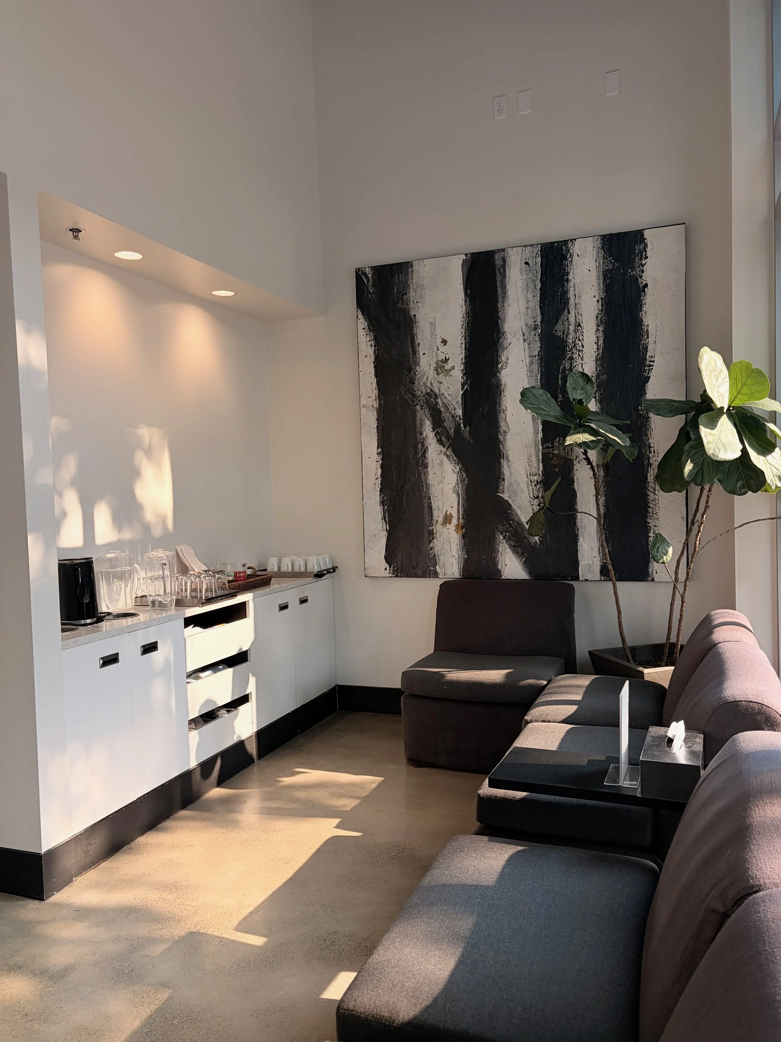 A lounge area with gray cushioned chairs, a large abstract black and white painting on the wall, a plant, and a white cabinet with glassware and coffee supplies, illuminated by natural sunlight.