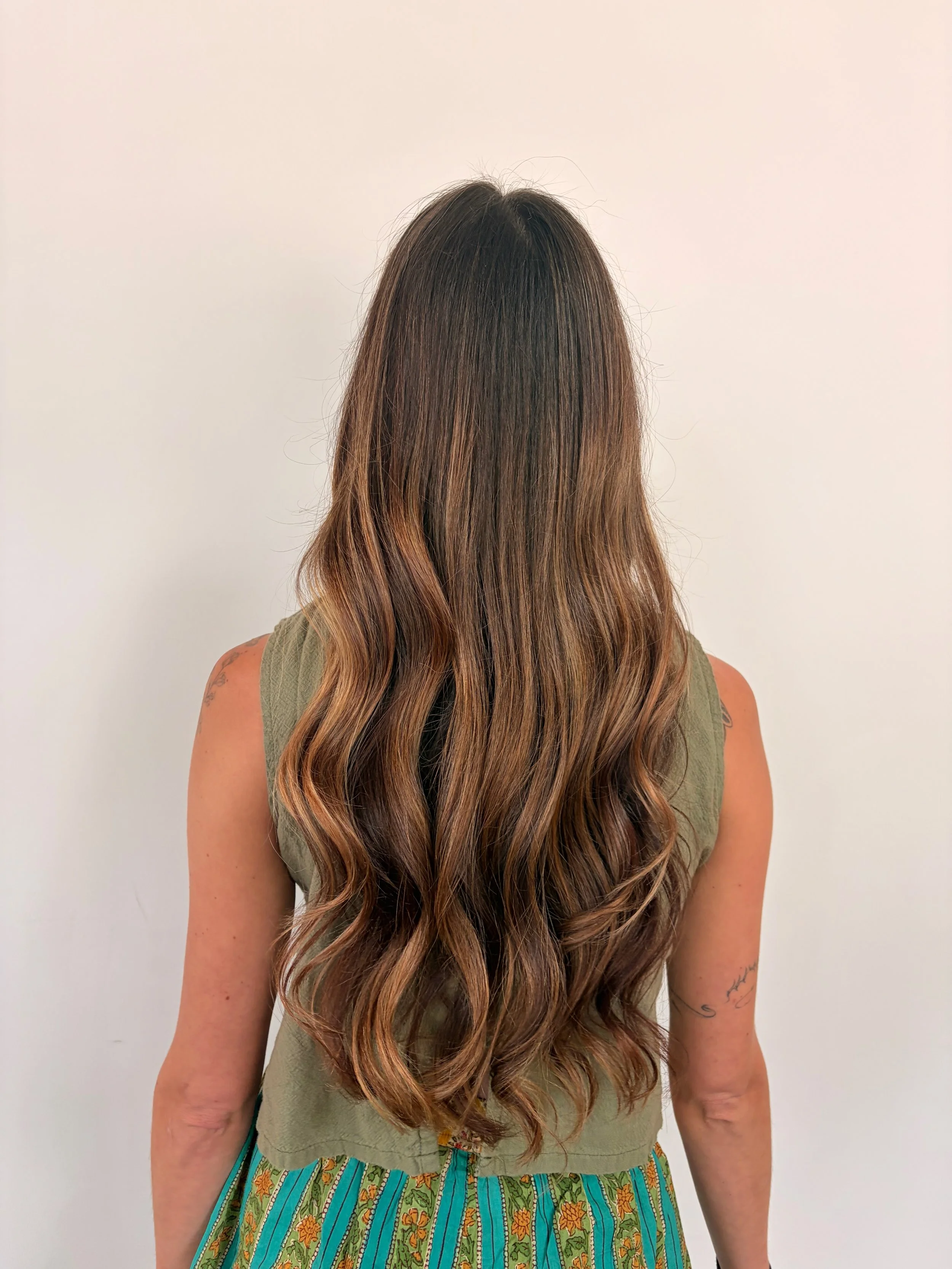 Back view of woman with long, wavy hair with caramel highlights, wearing a sleeveless olive green top and a patterned skirt with blue, yellow, and orange designs, standing against white background.