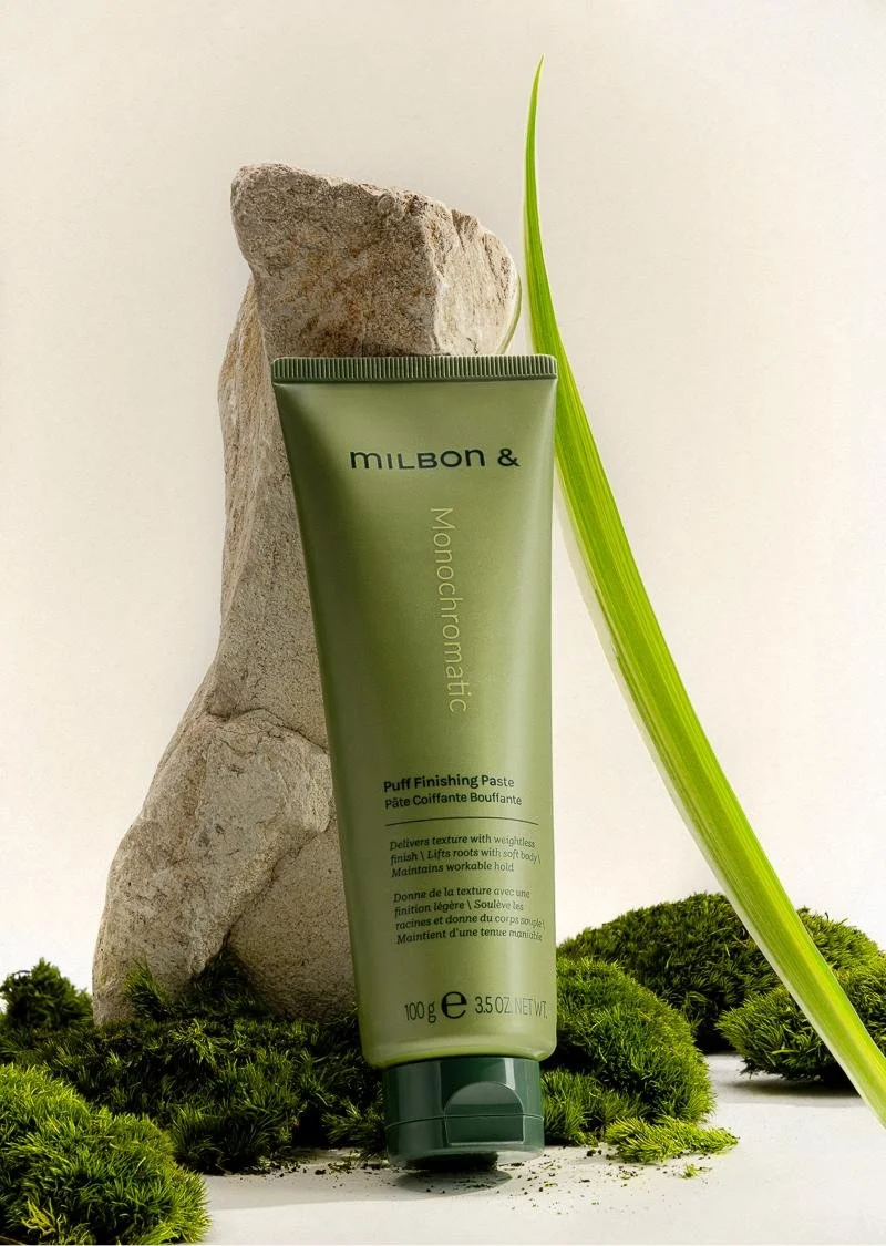 A tube of Milbon & Monochromatic Puff Finishing Paste is positioned standing upright outdoors on a bed of moss, leaning against a large stone, with a tall green grass blade beside it. The background is light and neutral.