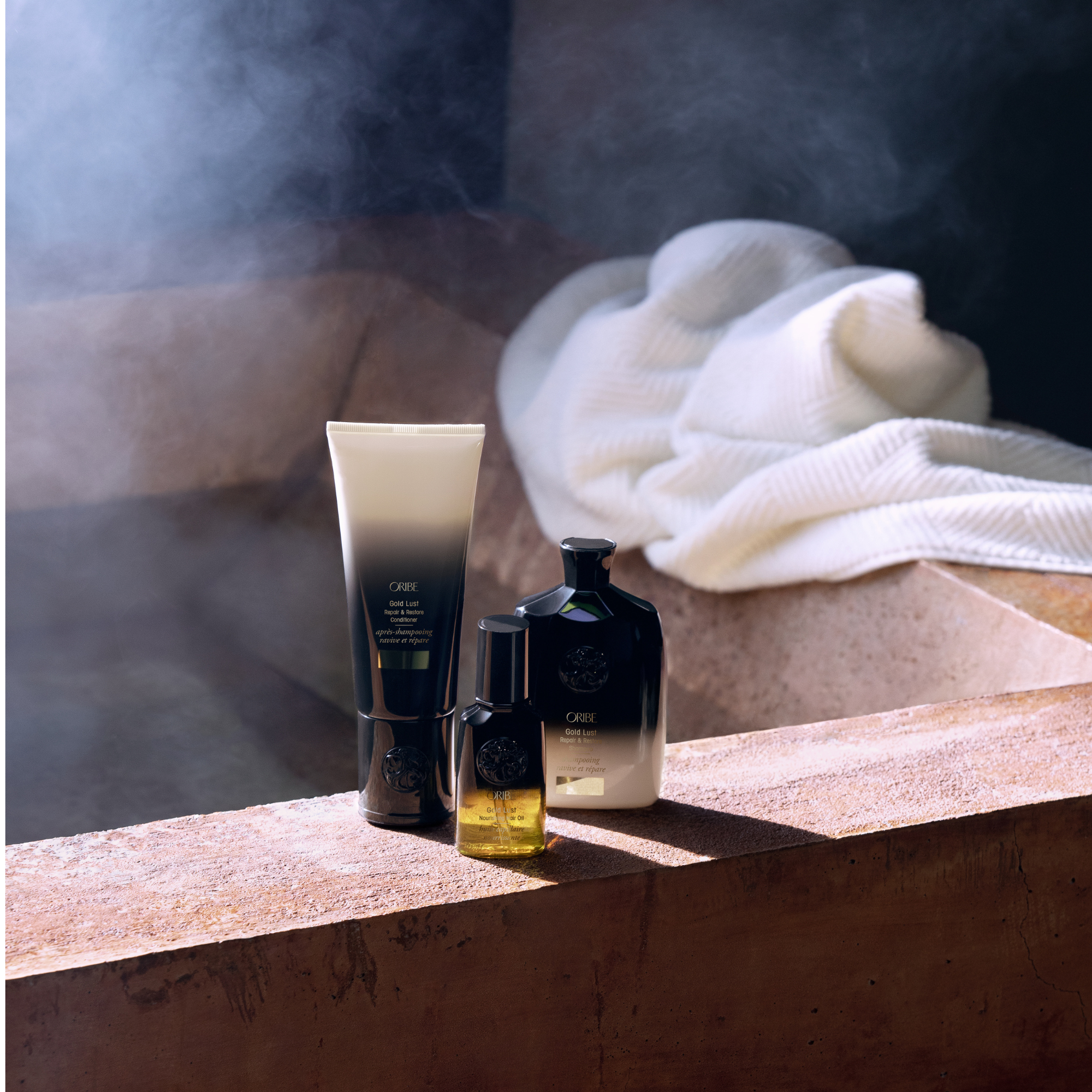 Collection of skin care products including a tube of ORIBE Gold Lust Repair & Restore Conditioner, a bottle of ORIBE Gold Lust Nourishing Hair Oil, and a few other bottles, all placed on a textured surface with a white cloth nearby.