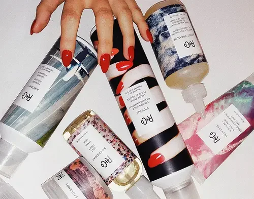 A hand with red nail polish holding a cylindrical bottle of R+Co hair product surrounded by five other bottles and boxes of Hair Care products with artwork and labels.