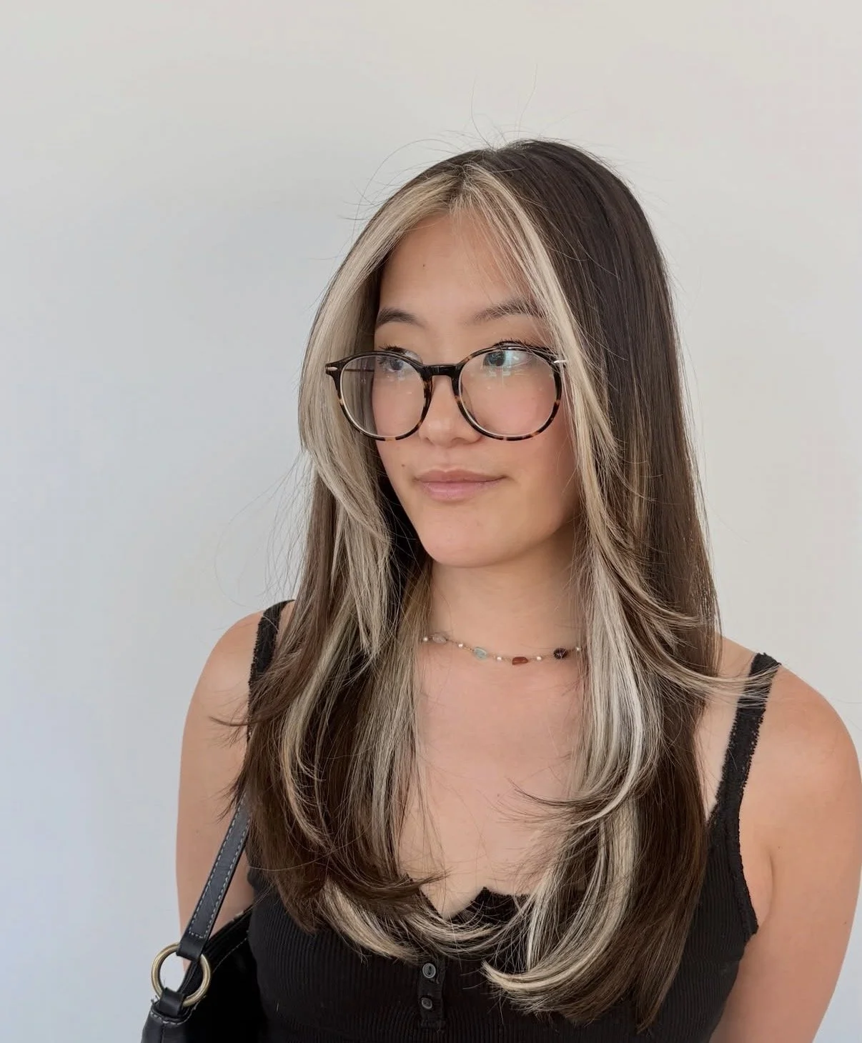 A woman with long, dark hair and blonde highlights wearing glasses, a black sleeveless top, and a beaded necklace, standing against a plain white background.