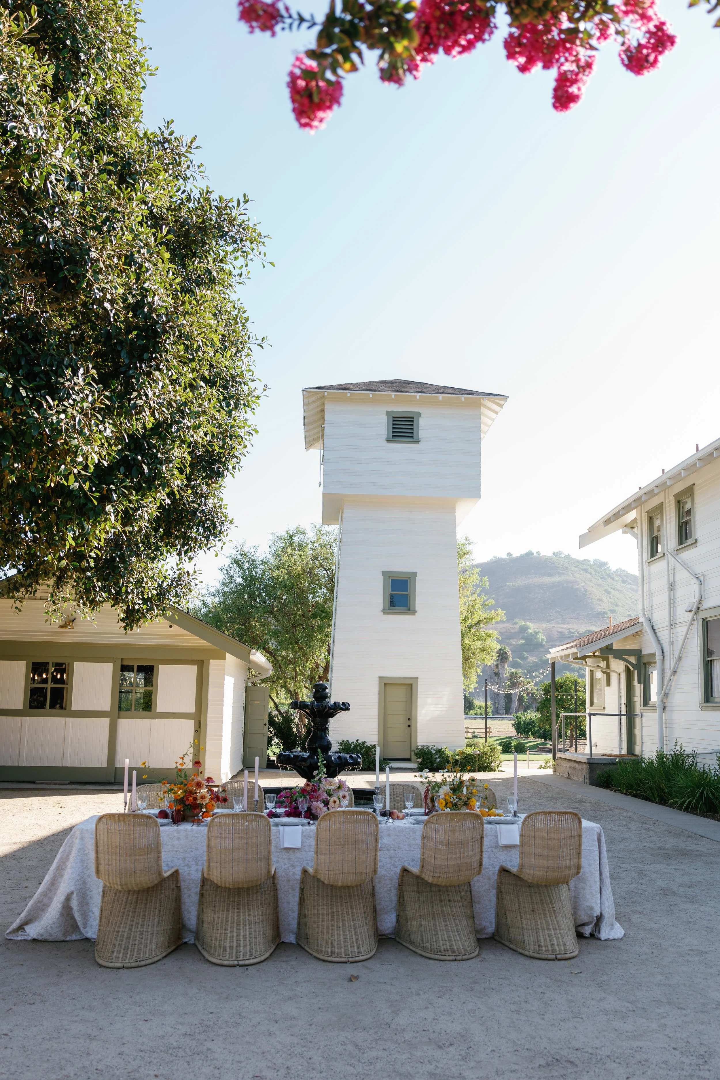 Outdoor event setup with a long rectangular table covered with a white tablecloth and decorated with vibrant floral arrangements. Wicker chairs are arranged around the table. In the background, there's a white building, a fountain, trees, and a hilly landscape under a clear blue sky.