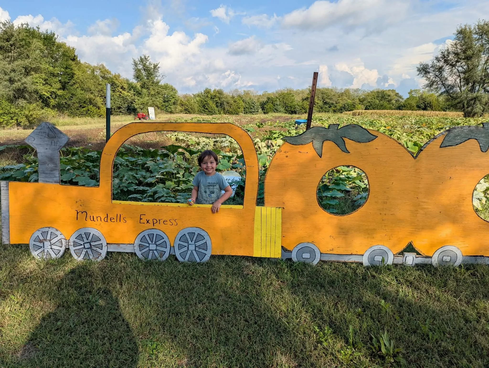 Child smiling inside a large, yellow, wooden train photo prop at a pumpkin patch or farm, with green fields and trees in the background under a partly cloudy sky.