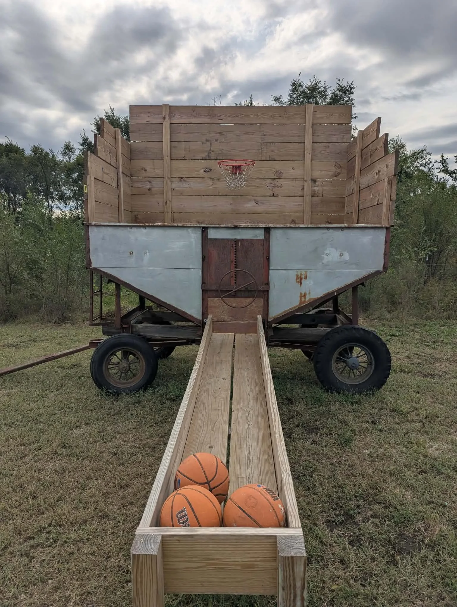 A backyard basketball hoop attached to a wooden privacy fence, with a wooden ramp leading to the hoop and three basketballs in a wooden box, set on grassy ground under cloudy sky.