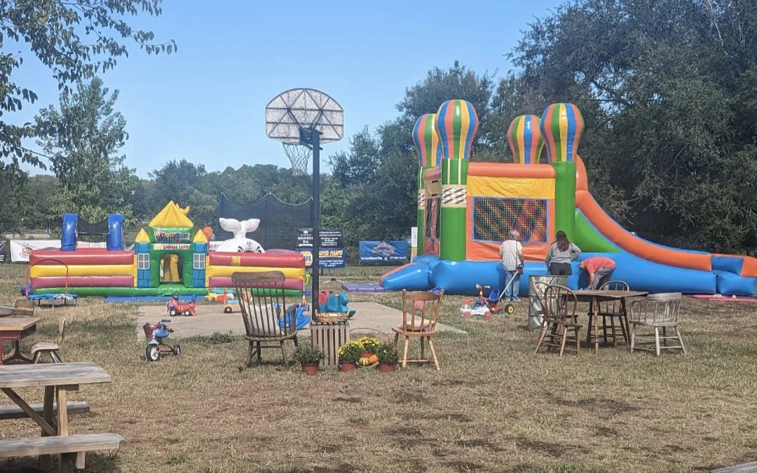 Outdoor children's play area with inflatable bounce houses, a basketball hoop, and various outdoor furniture, including chairs and tables, in a grassy area surrounded by trees.