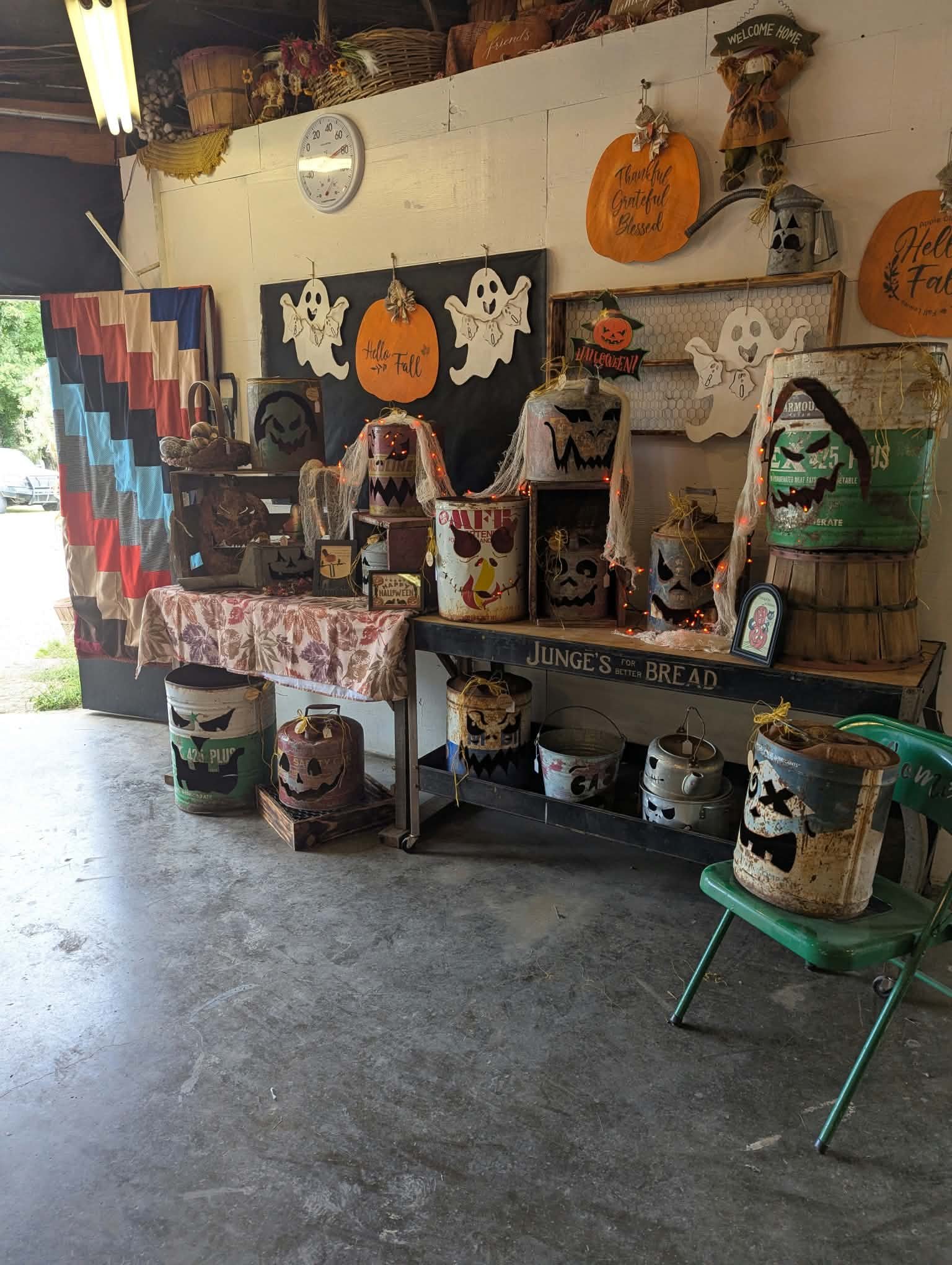 Part of a fall-themed display with Halloween decorations, painted and metal buckets with pumpkin faces, ghost cutouts, and signs that say "Hello Fall," "Thankful, Grateful, Blessed", and "Welcome Home." There is a table with a floral tablecloth, and 
