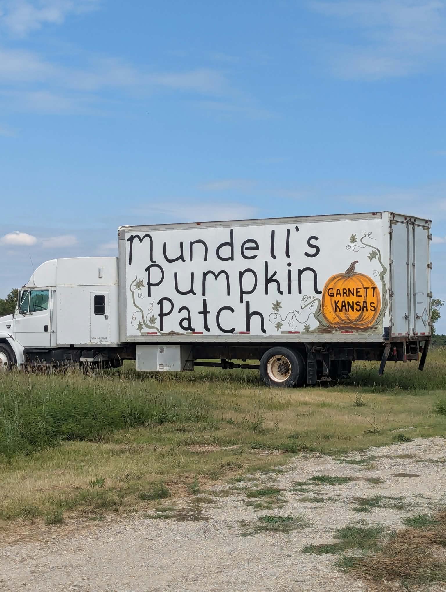 A white truck with a large sign on its side that reads 'Mundell's Pumpkin Patch' and features a pumpkin graphic with 'Garnett Kansas' written inside, parked on a grassy area under a blue sky with scattered clouds.