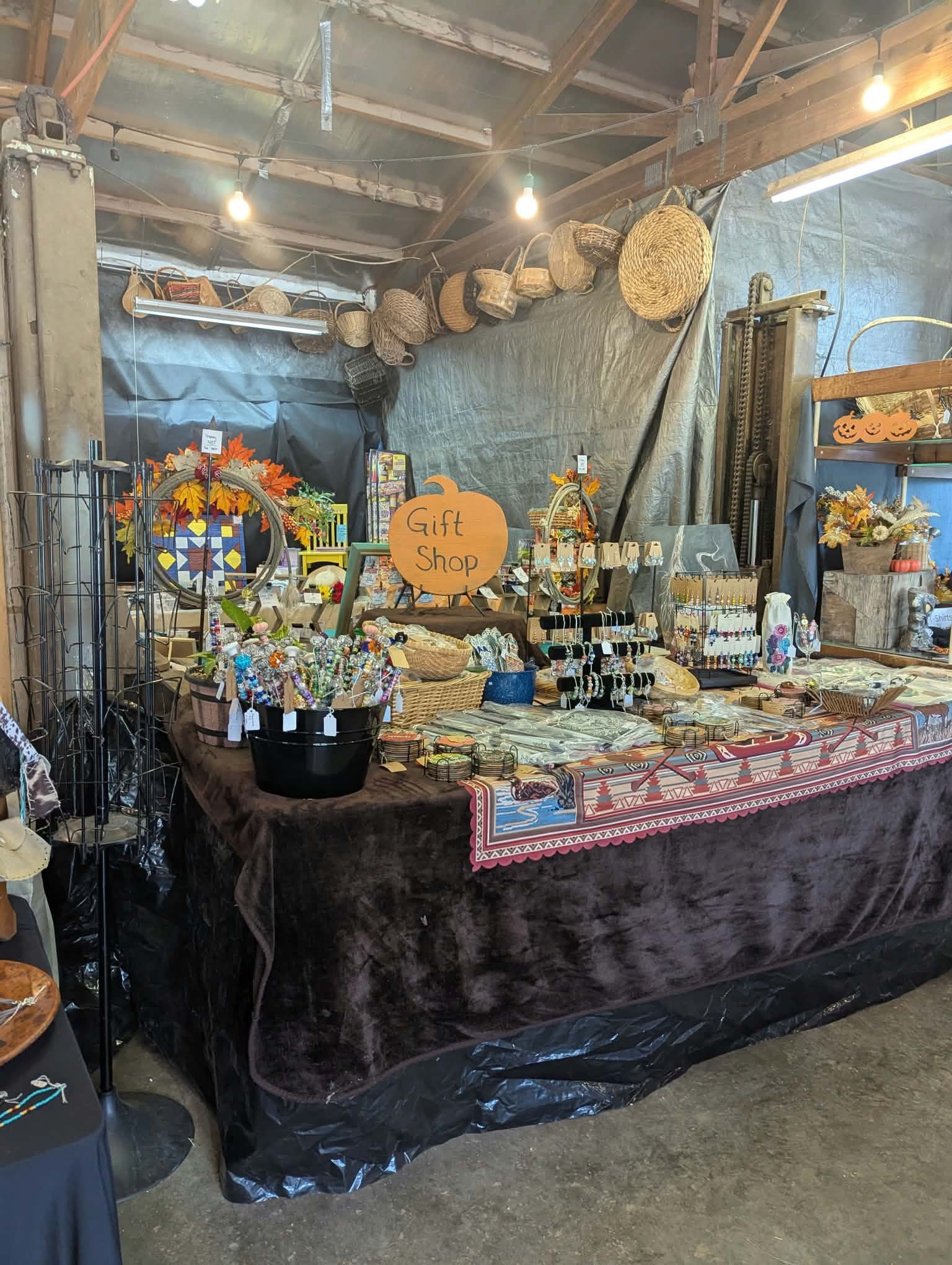 A gift shop booth displaying jewelry, ornaments, and crafts with a pumpkin sign and fall decorations, set up inside a barn with wicker baskets hanging overhead.