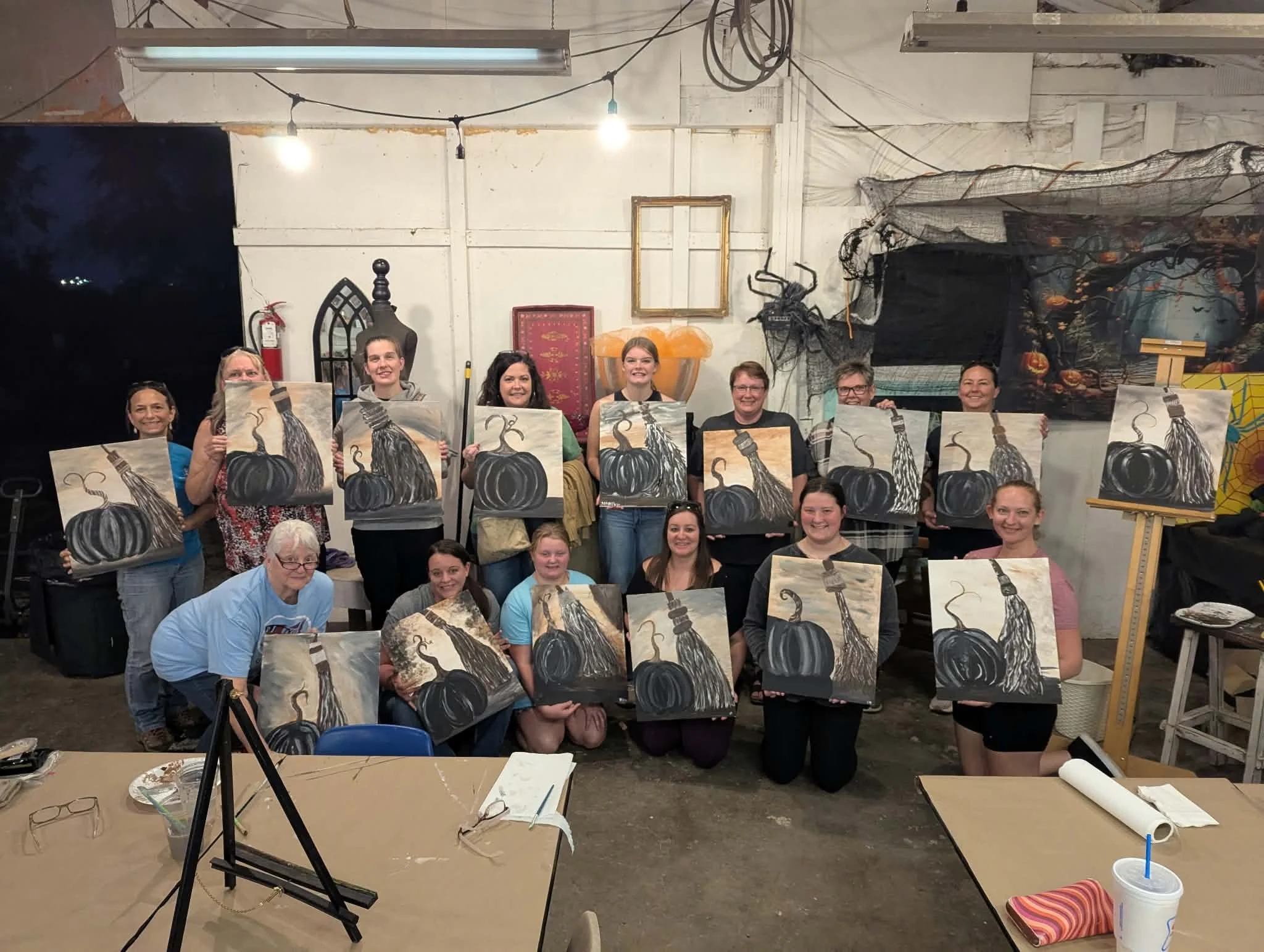 A group of people holding paintings of black pumpkins with curly stems in an art studio. The studio has a white concrete wall with Halloween decorations, including black netting and a framed picture of a pumpkin scene. There are art supplies and equipment on tables and in the background.
