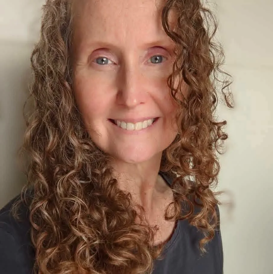 Close-up of a woman with curly brown hair, blue eyes, smiling, wearing a dark top.