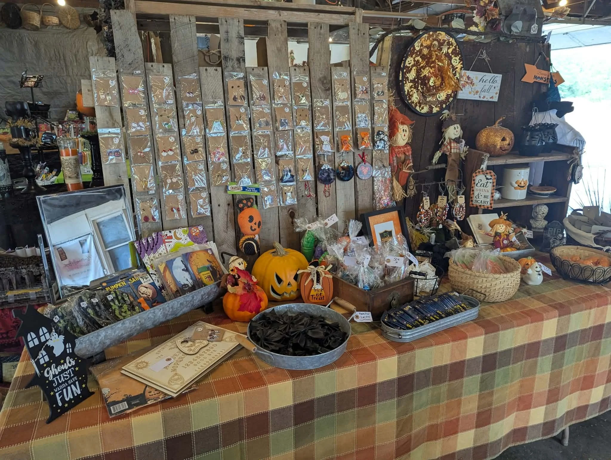 A display of Halloween-themed decorations and accessories at a market stall. Items include pumpkin decorations, spooky signs, and craft jewelry on a wooden board. The table has a checkered orange and green tablecloth, and various other decorative ite