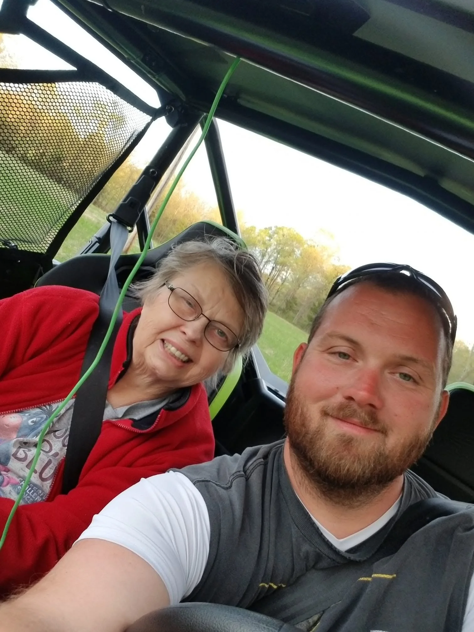 A selfie of a smiling elderly woman with glasses and a man inside a vehicle, with a green landscape and trees visible through the window in the background.