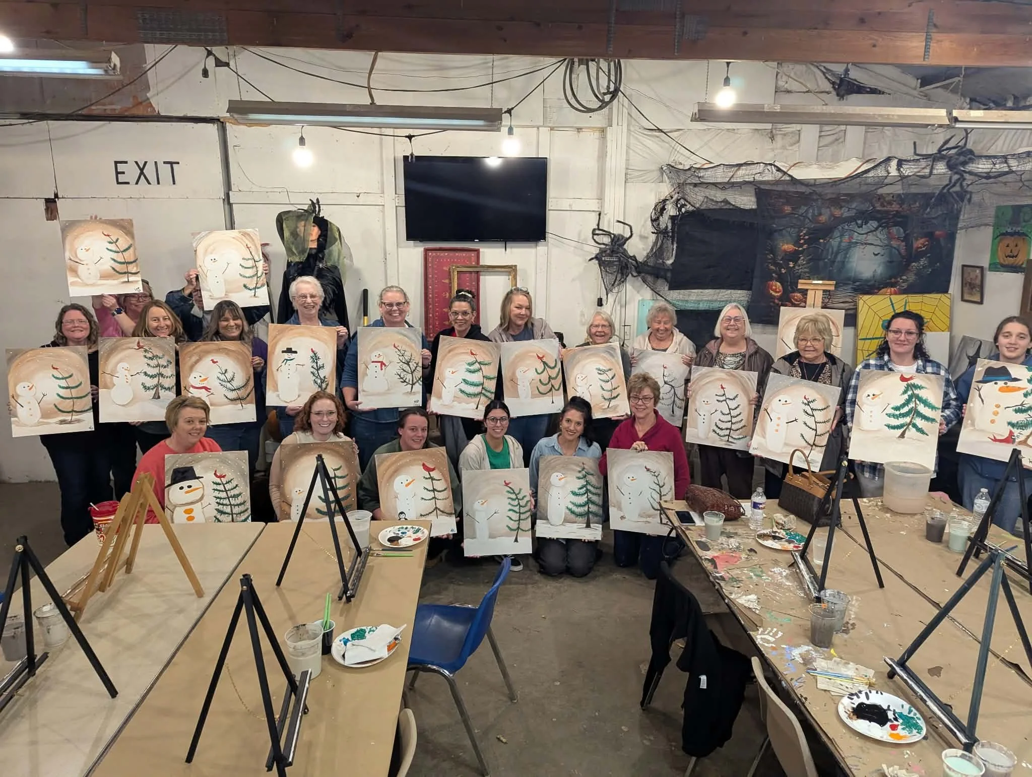 Group of people holding up paintings of snowmen and Christmas trees, with painting supplies and easels in the foreground, in a room decorated for Halloween with spooky web and pumpkin decorations.