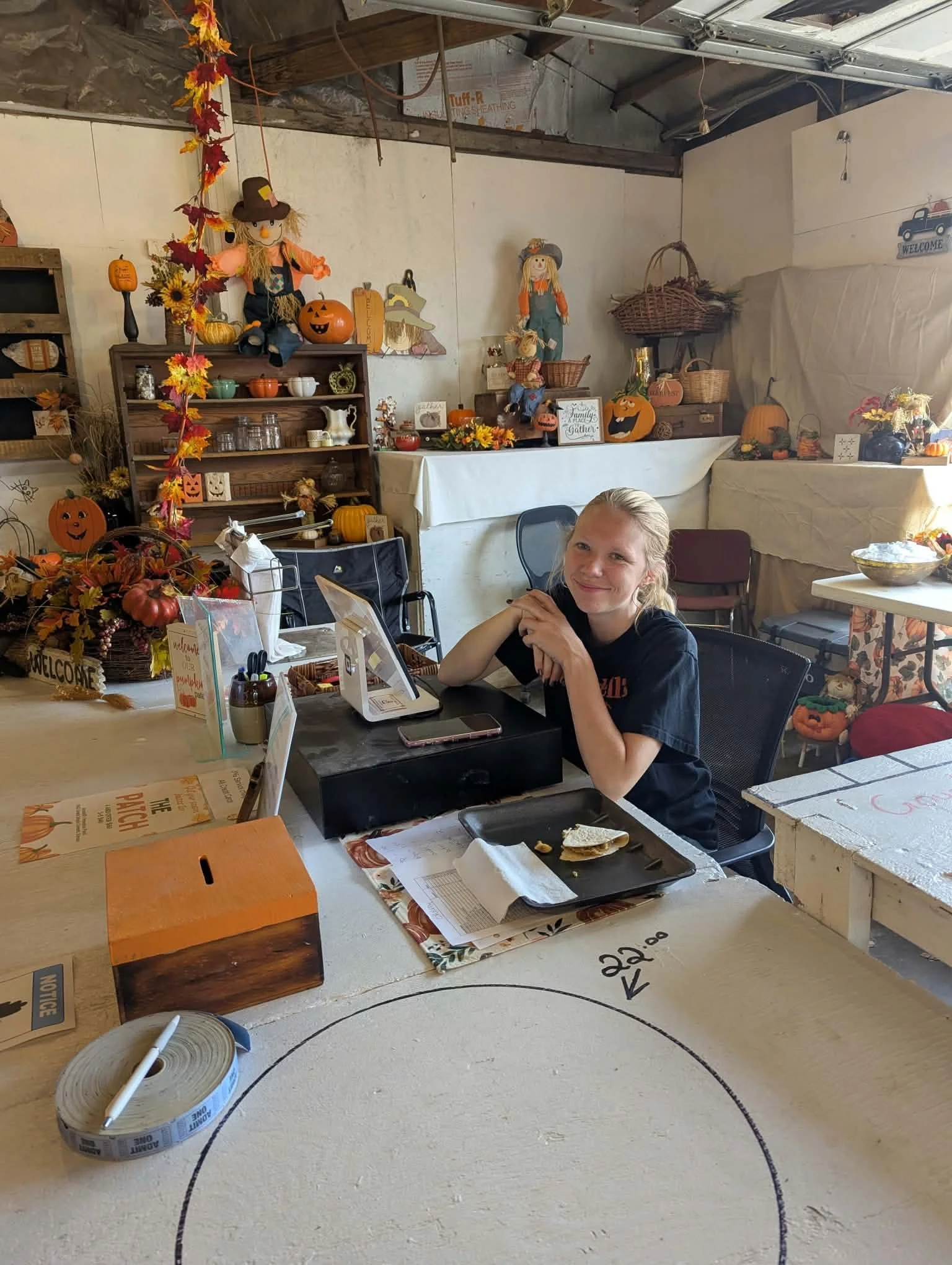 A woman sitting at a table in a room decorated for fall, with autumn-themed decorations, pumpkins, scarecrows, and harvest arrangements visible in the background.