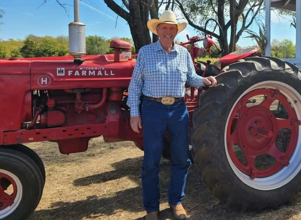 An elderly man in cowboy attire, wearing a cowboy hat, plaid shirt, jeans, and boots, standing next to a red McCormick Farmall tractor outdoors on a farm with trees in the background.