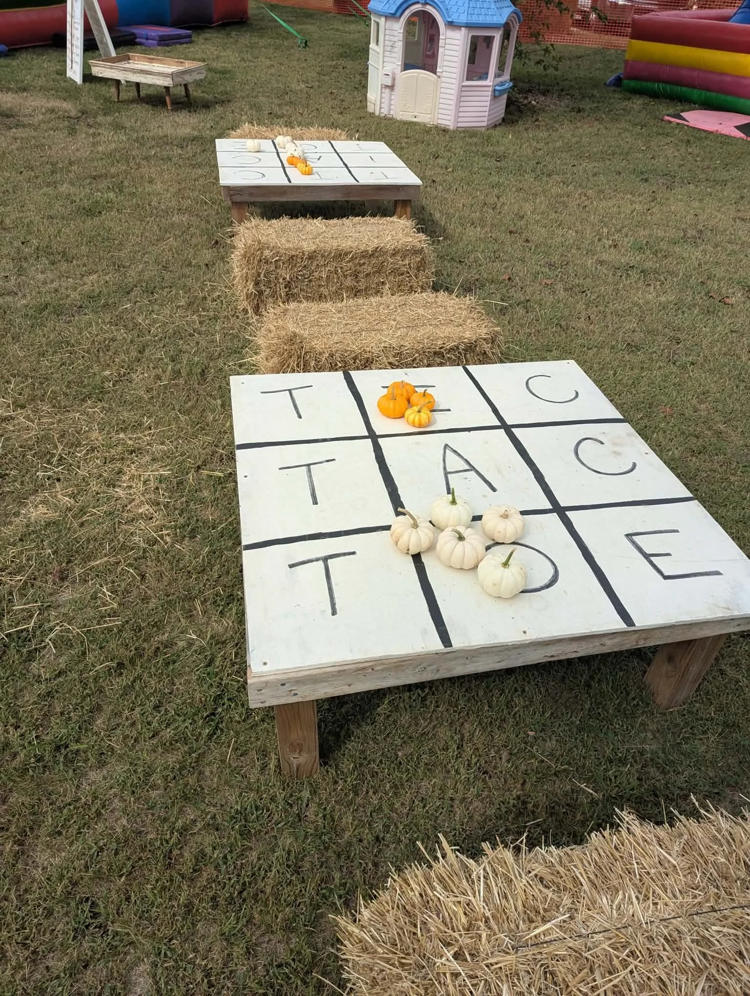 Outdoor scene with hay bales, a small plastic house, and a games table with pumpkins and gourds, in a grassy area with orange fencing.