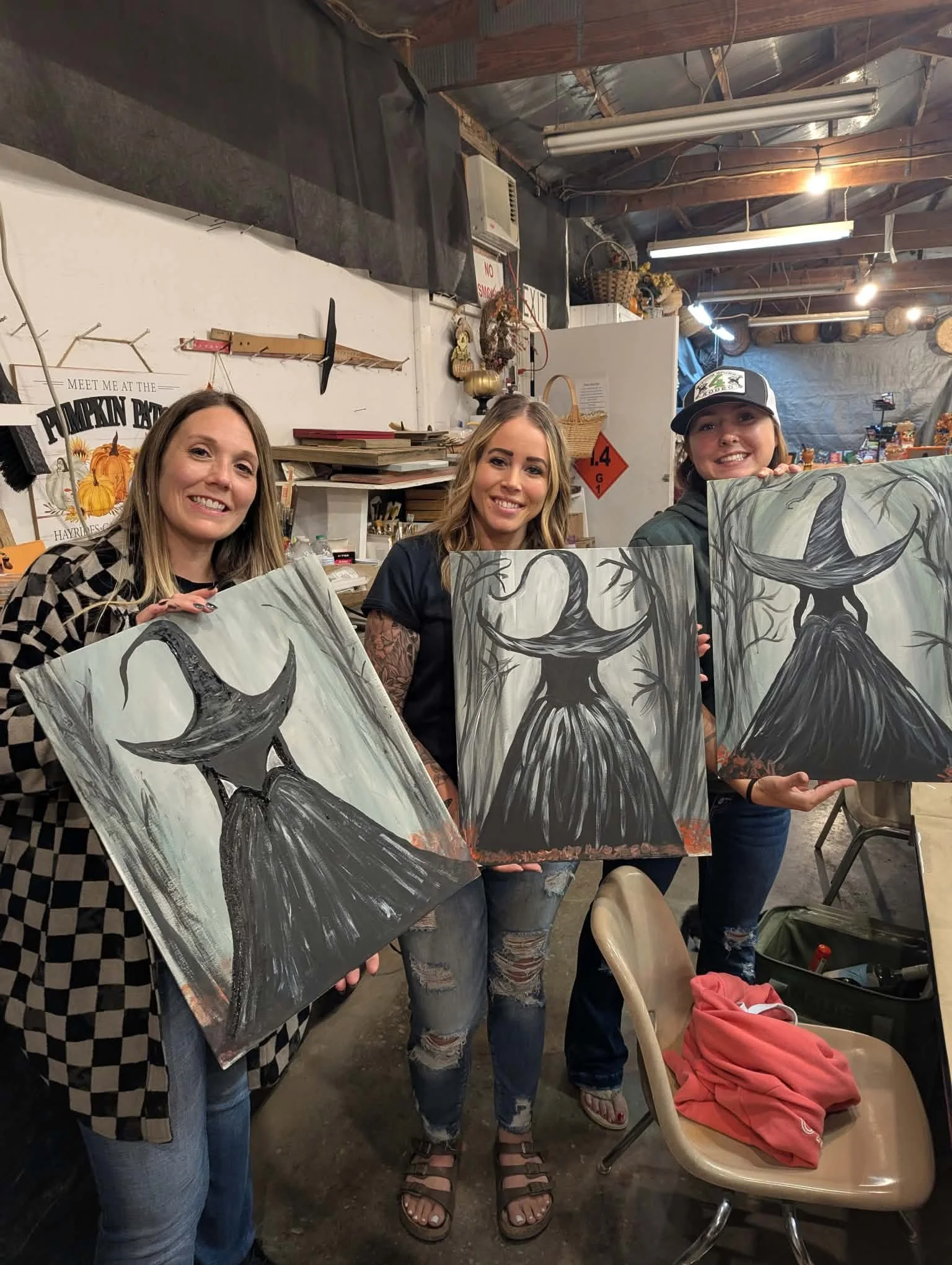 Three women holding paintings of black, fairy-tale style trees with large, twisting branches, inside a rustic art studio or workshop.