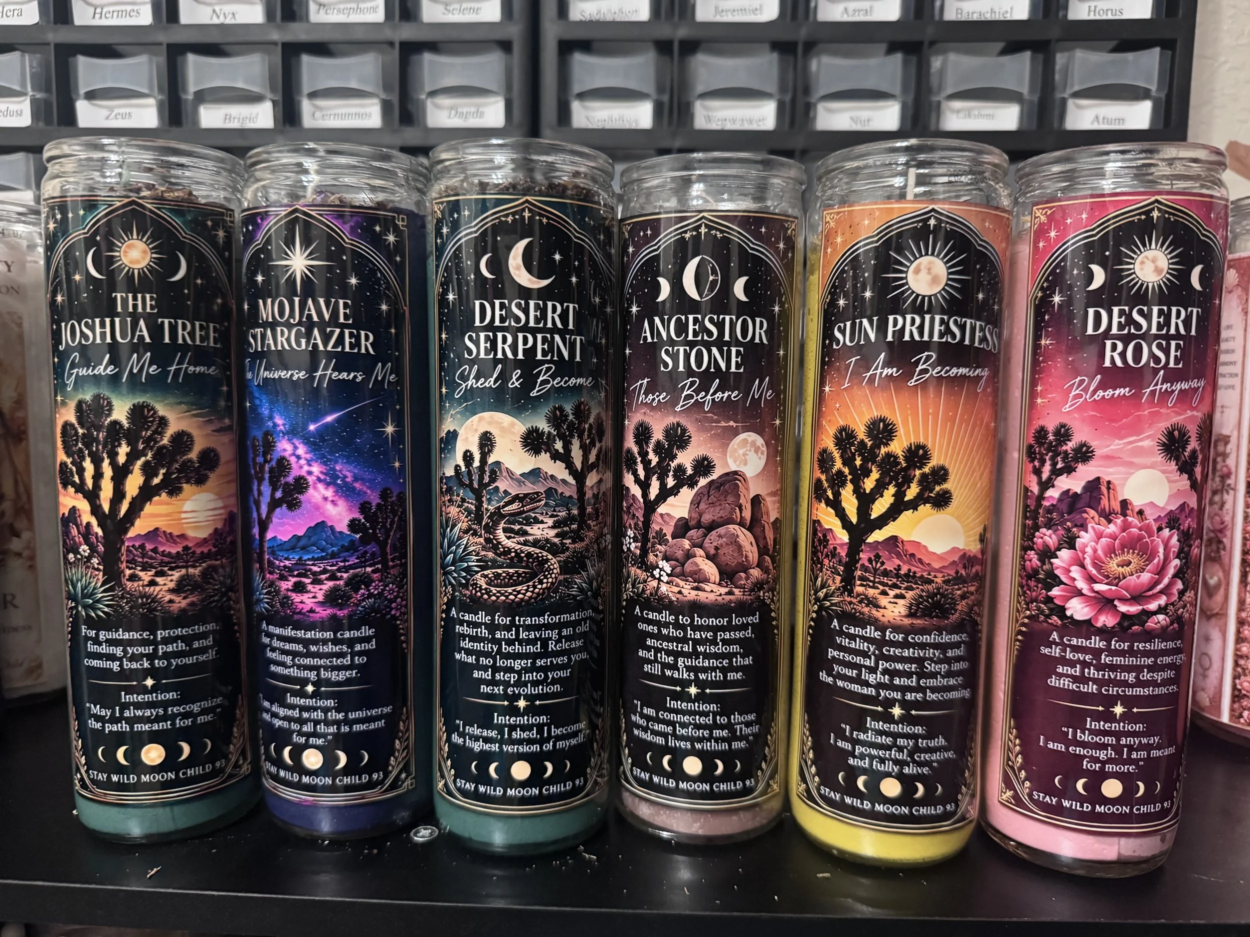 A row of candles with colorful desert-themed labels, featuring celestial and desert landscape designs, each with a different poetic or inspirational message.