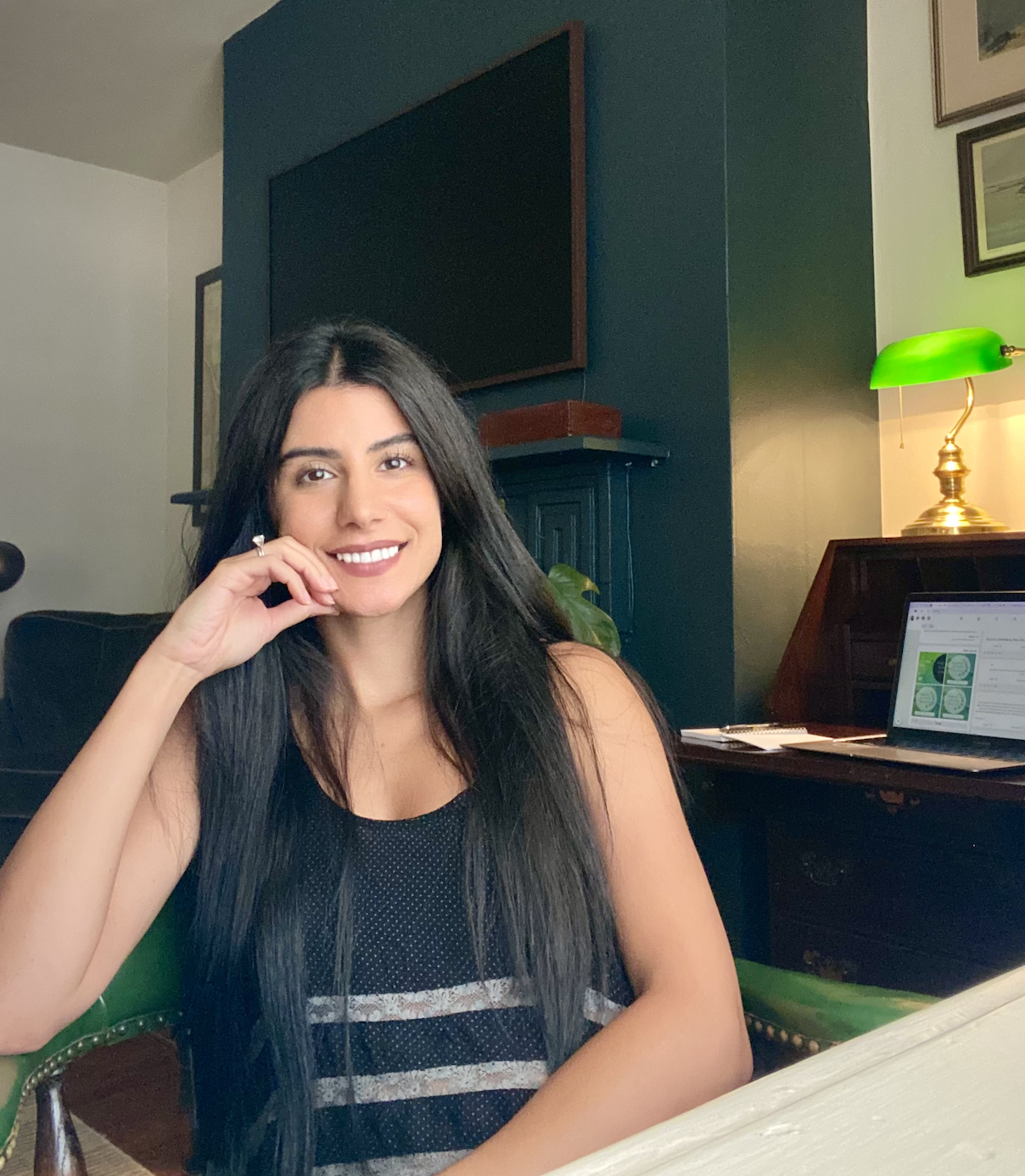 A woman with long dark hair smiling and sitting at a table in a room with green walls, a lamp with a green shade, and a laptop open to a webpage.