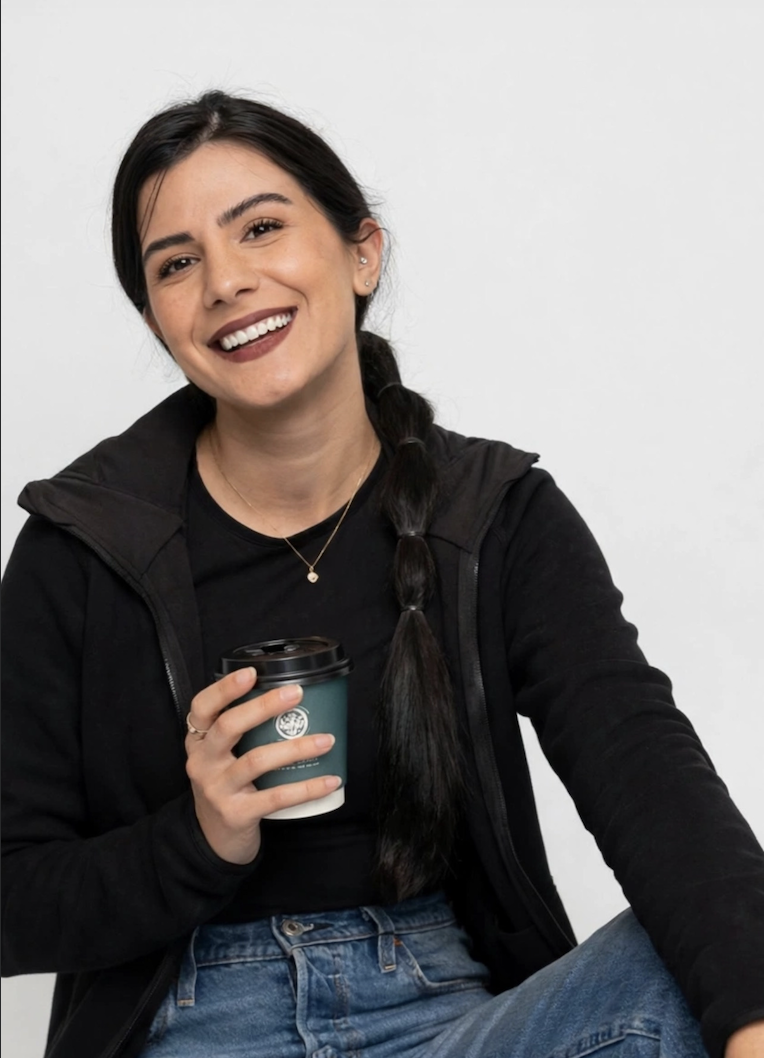 A woman with dark hair in a braid, smiling and holding a to-go coffee cup, sitting against a plain white wall.