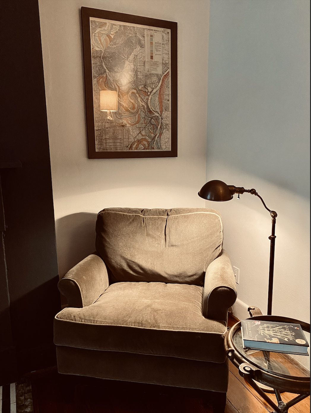 A cozy living room corner with a beige upholstered armchair, a black floor lamp standing next to it, a small circular glass-top table with a book on it, and a framed map hanging on the white wall behind the chair.