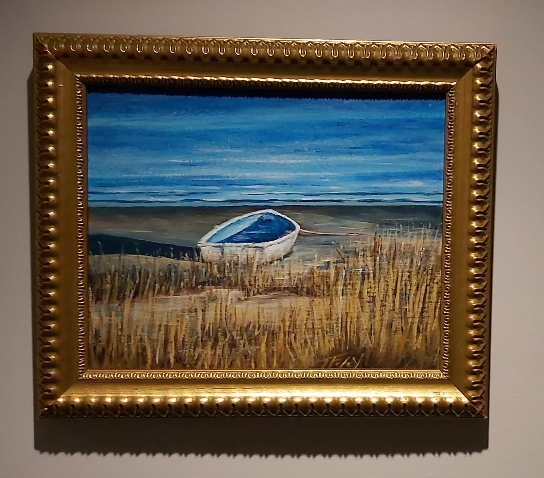 A framed painting of a boat resting on a sandy shore with a calm body of water and a clear blue sky in the background.