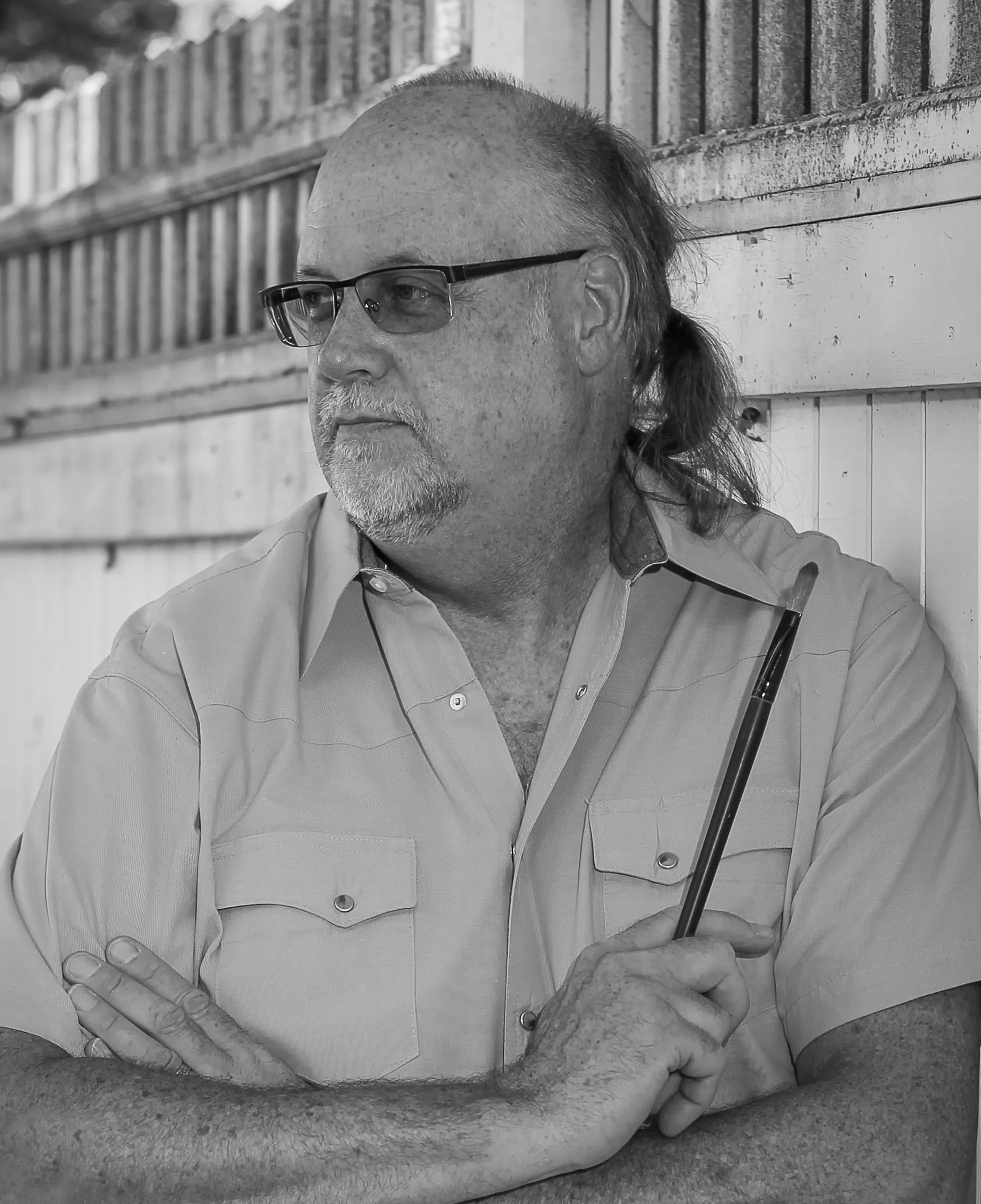 A person with glasses and a partially shaved head, sitting outdoors against a wooden fence, holding a pen, with arms crossed and leaning on a surface.