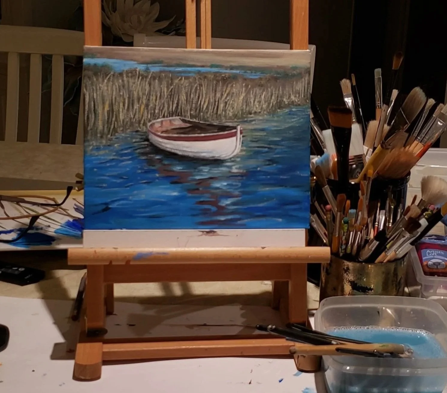 A painting on an easel depicting a boat floating on blue water near a shoreline with tall grass and a distant sky.