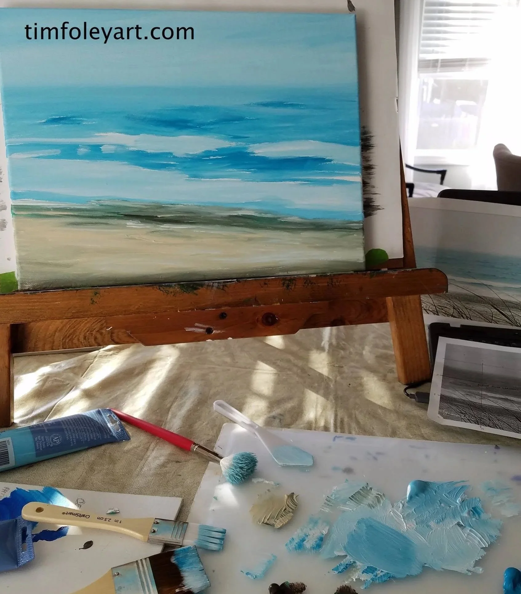 A painting of a seascape with blue sky, white clouds, and sandy beach, placed on an easel in a bright room. Painting supplies including brushes, paint tubes, and palette knives are scattered on a table in front of the easel.