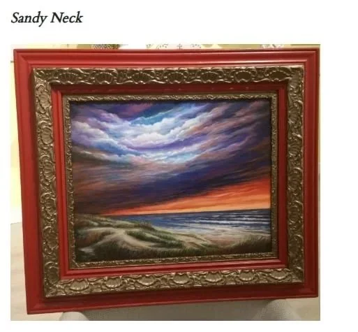 A framed landscape painting of a dramatic sky with dark clouds and a sunset over a beach with grassy dunes.