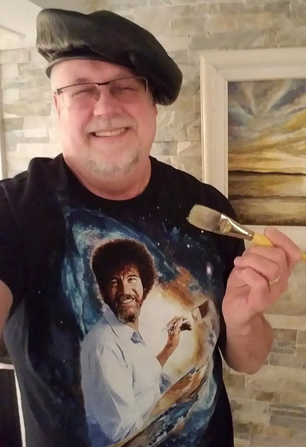 A man with glasses and a beret smiling while holding a paintbrush in front of a framed landscape painting on a brick wall.