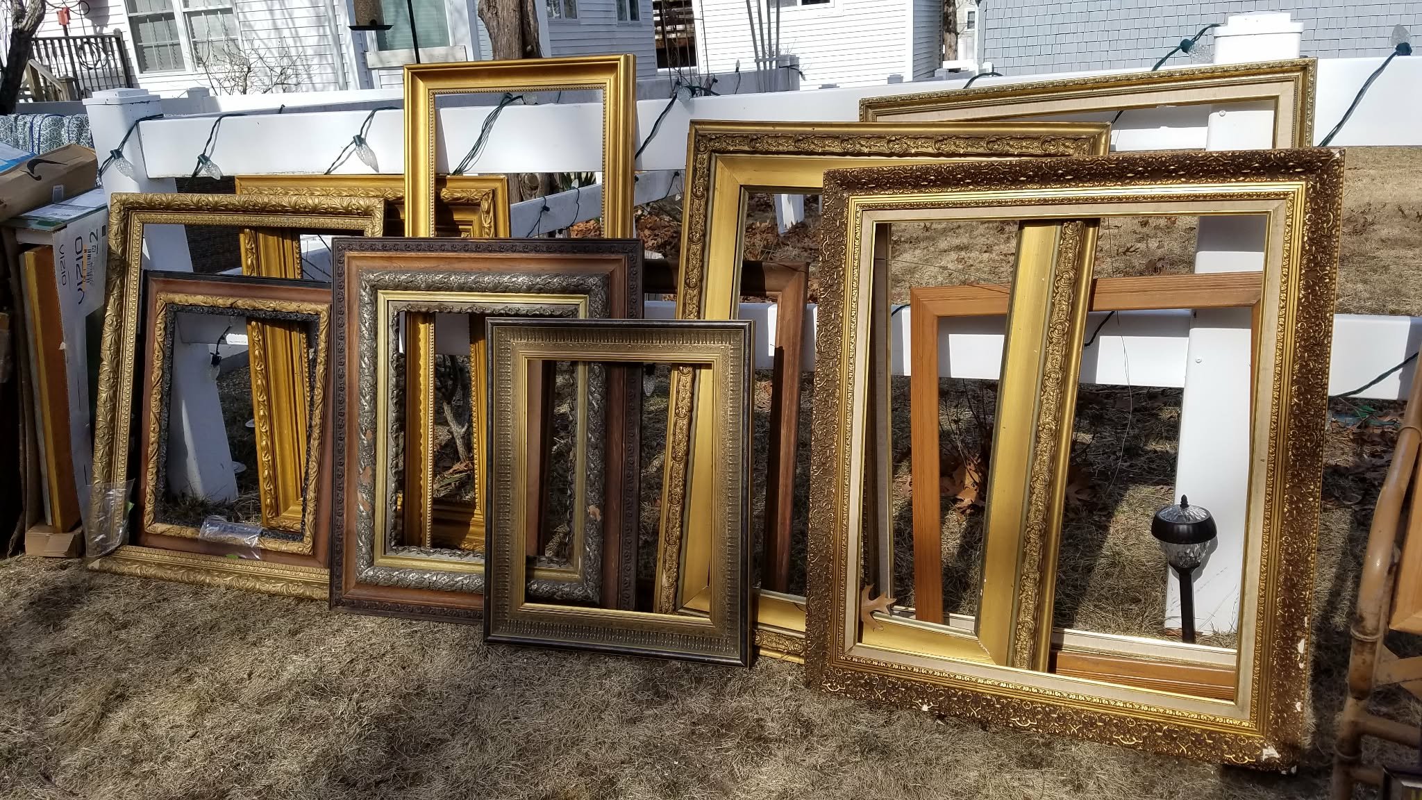 Multiple empty picture frames of various sizes and colors leaning against a white fence outdoors, with string lights hanging on the fence and a lamp at the bottom right.
