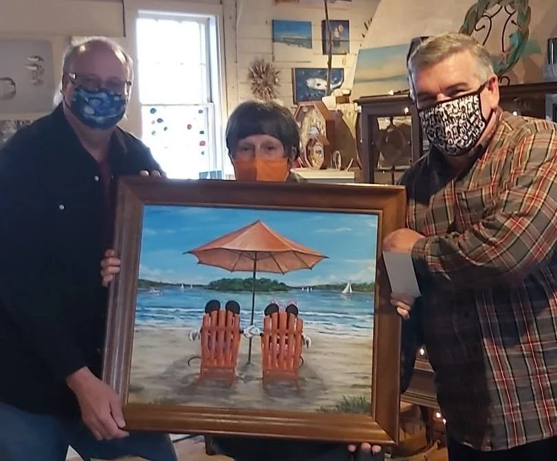 Three people holding a framed painting of a beach scene with two Adirondack chairs under an umbrella, by the water with sailboats.