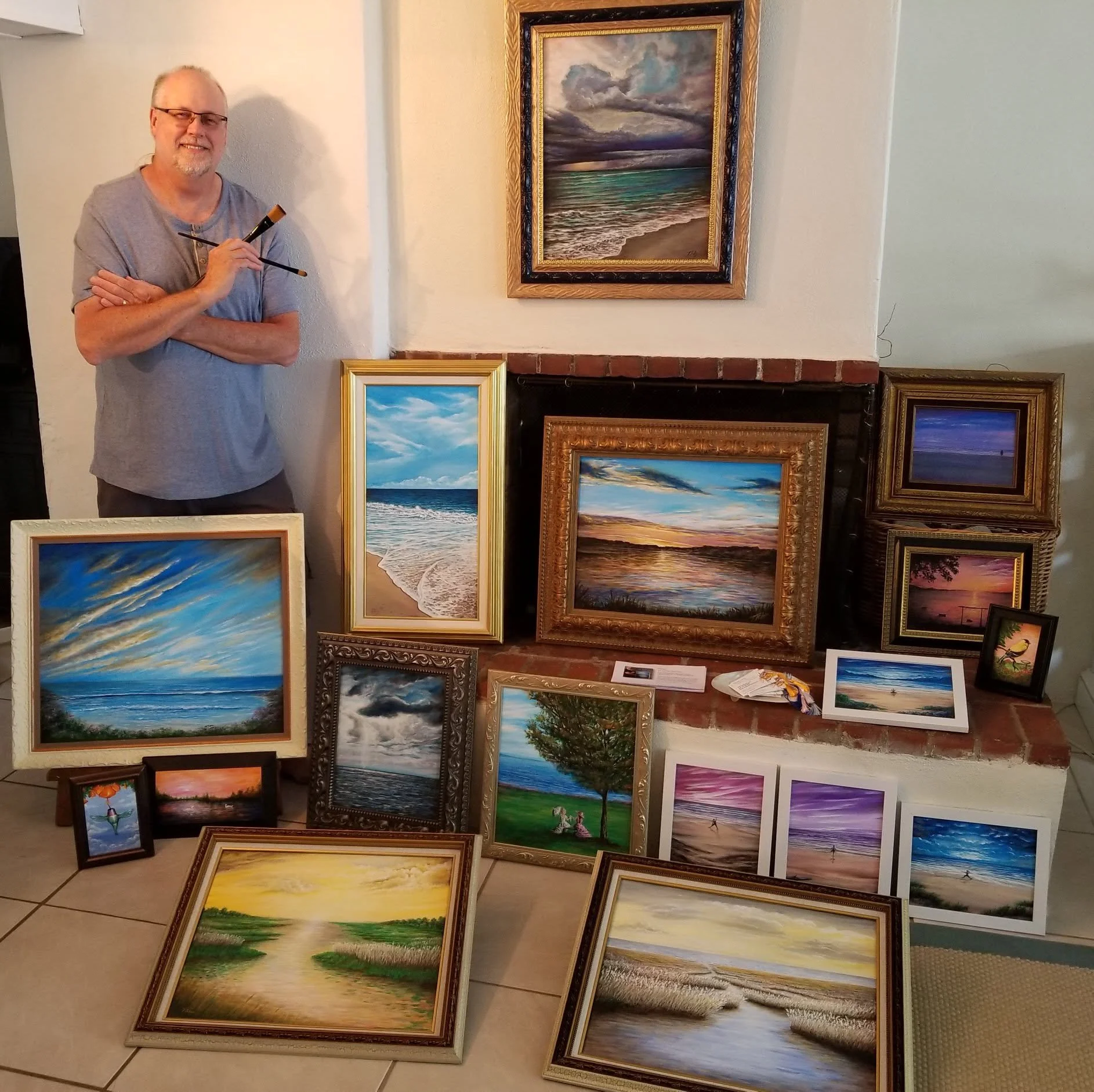 A man with glasses and a gray shirt standing in front of a display of various landscape paintings, including ocean scenes, trees, and sunset views.