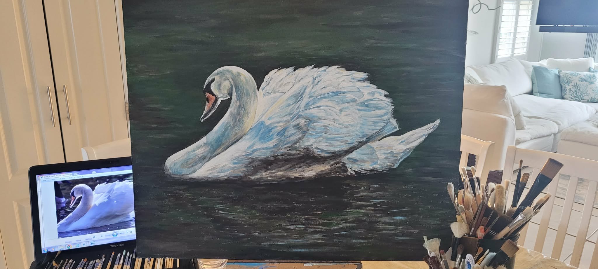 A painting of a white swan on dark water, placed on an easel in a living room with white furniture and light-filled windows.