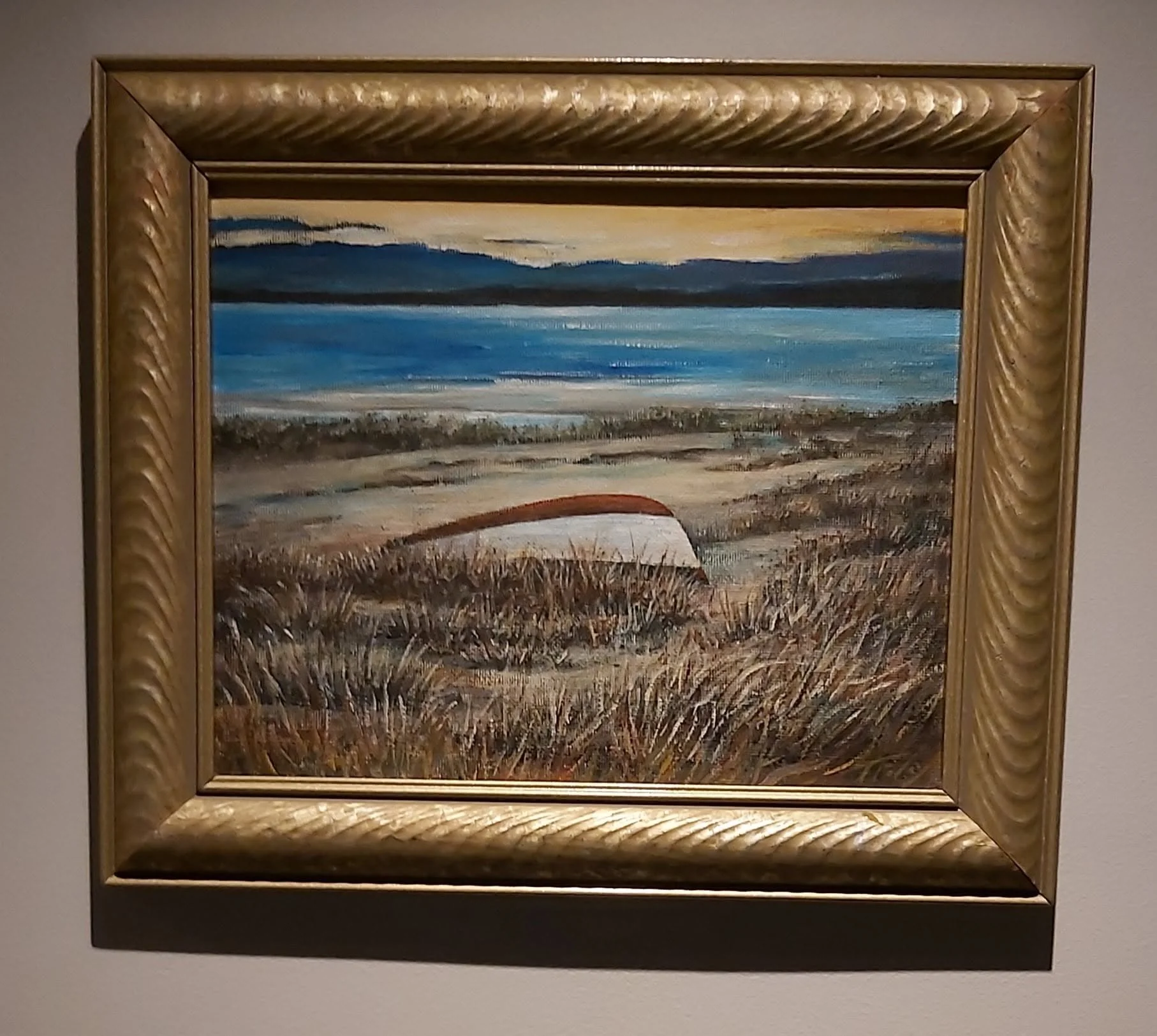 Oil painting of a beach scene with water, mountains, and sky, framed in ornate gold.