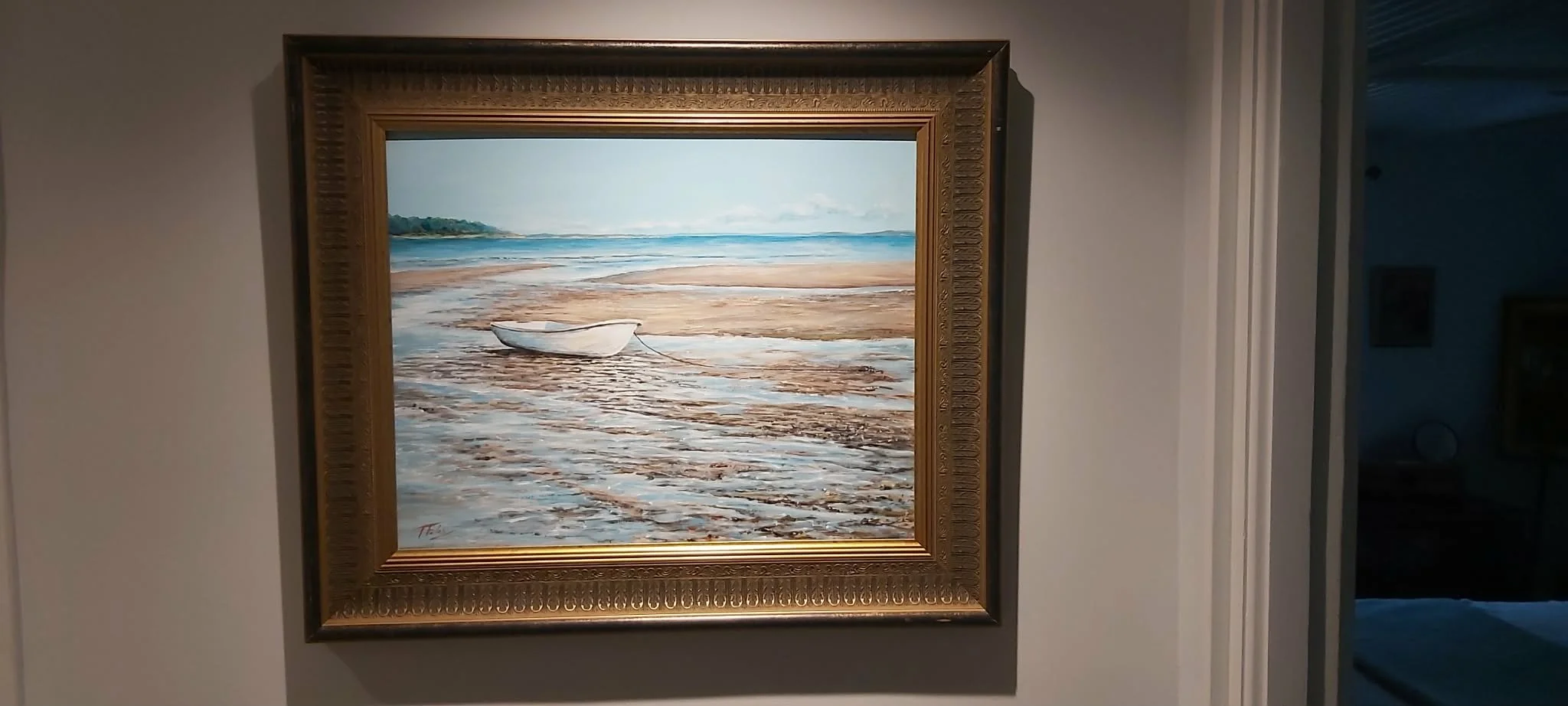 Painting of a boat on a beach with water and sky in the background, framed in an ornate gold frame hanging on a white wall.