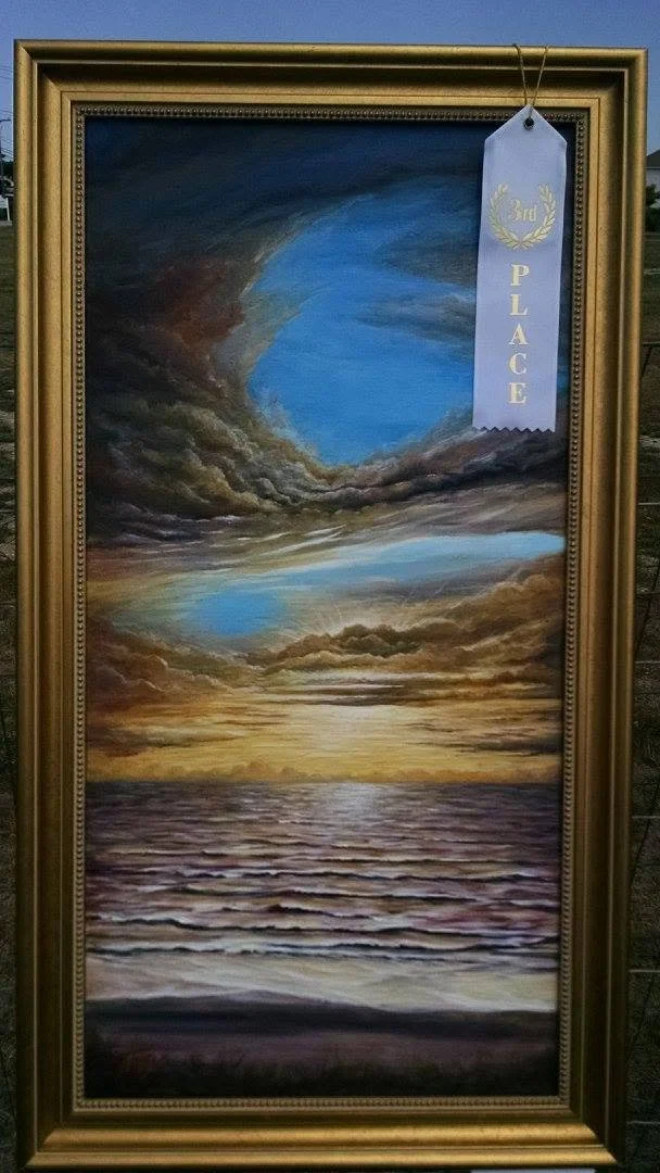 A painting of a sunset over the ocean with a dramatic sky featuring clouds, with a blue sky opening through the clouds, framed in a gold ornate frame and a blue ribbon indicating third place.