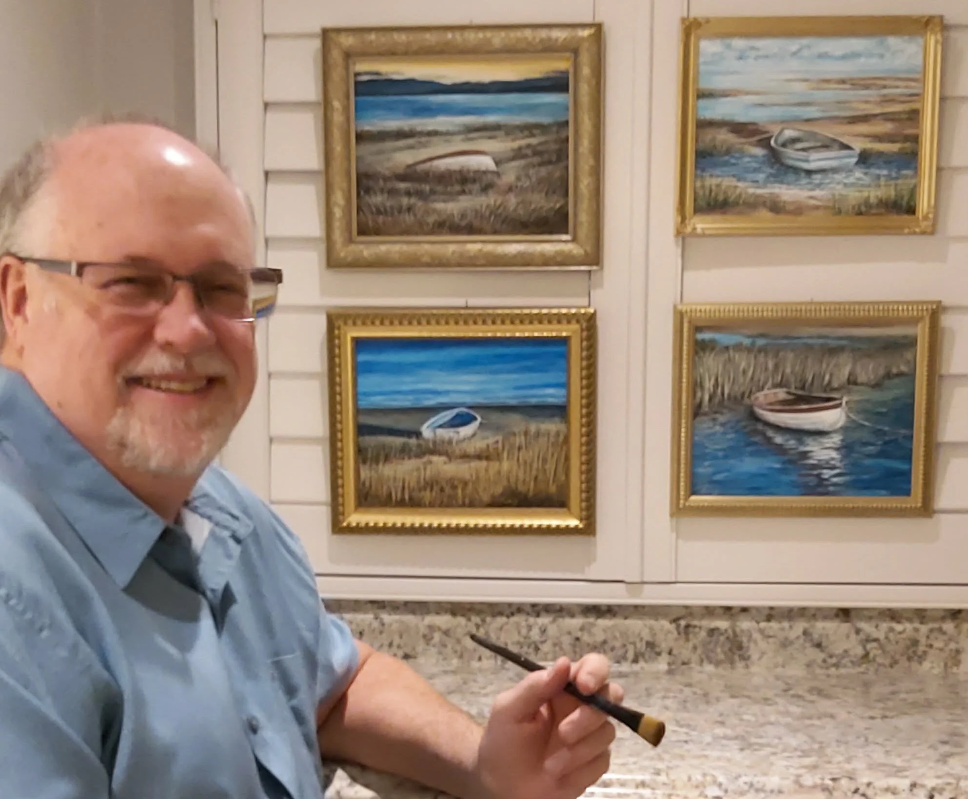 A smiling man with glasses and a beard holding a soldering iron in a room with framed landscape paintings on white panel walls.